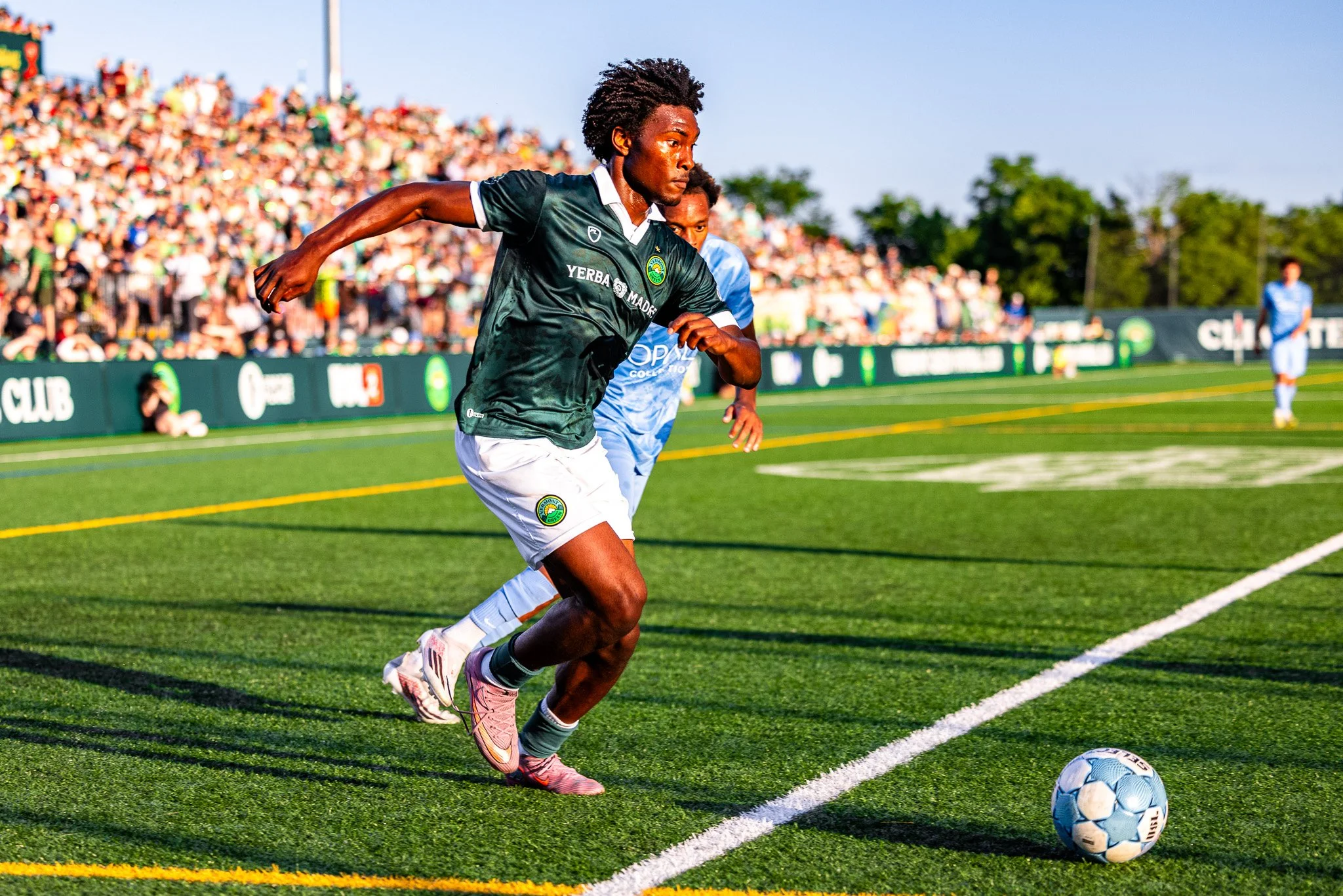 A soccer player with dark skin and curly hair running on a field with a ball in front of him, wearing a green jersey and white shorts, during a match with a crowd in the background.