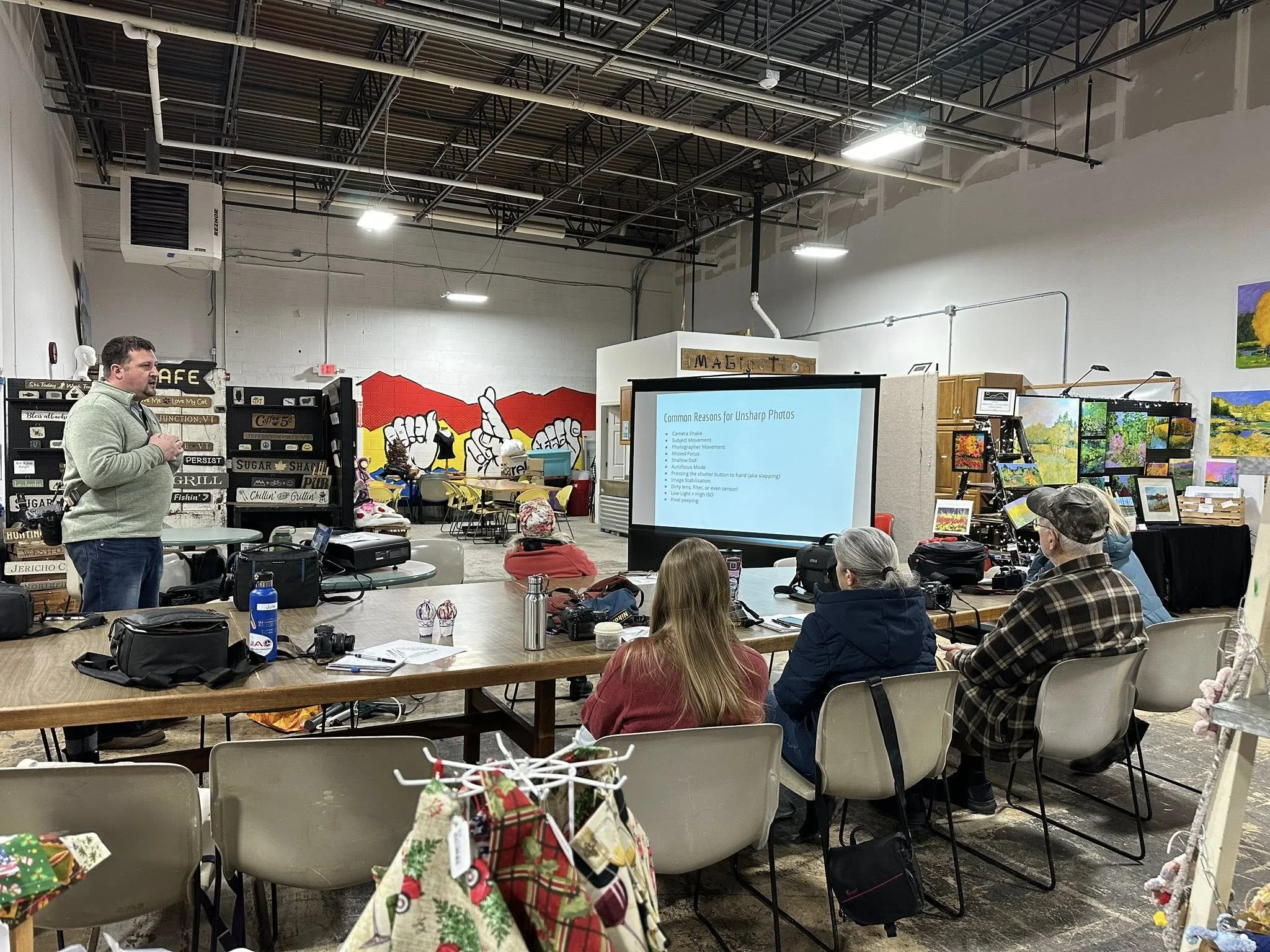 A man giving a presentation to a seated audience in a gallery or art space, with art displays and paintings in the background.
