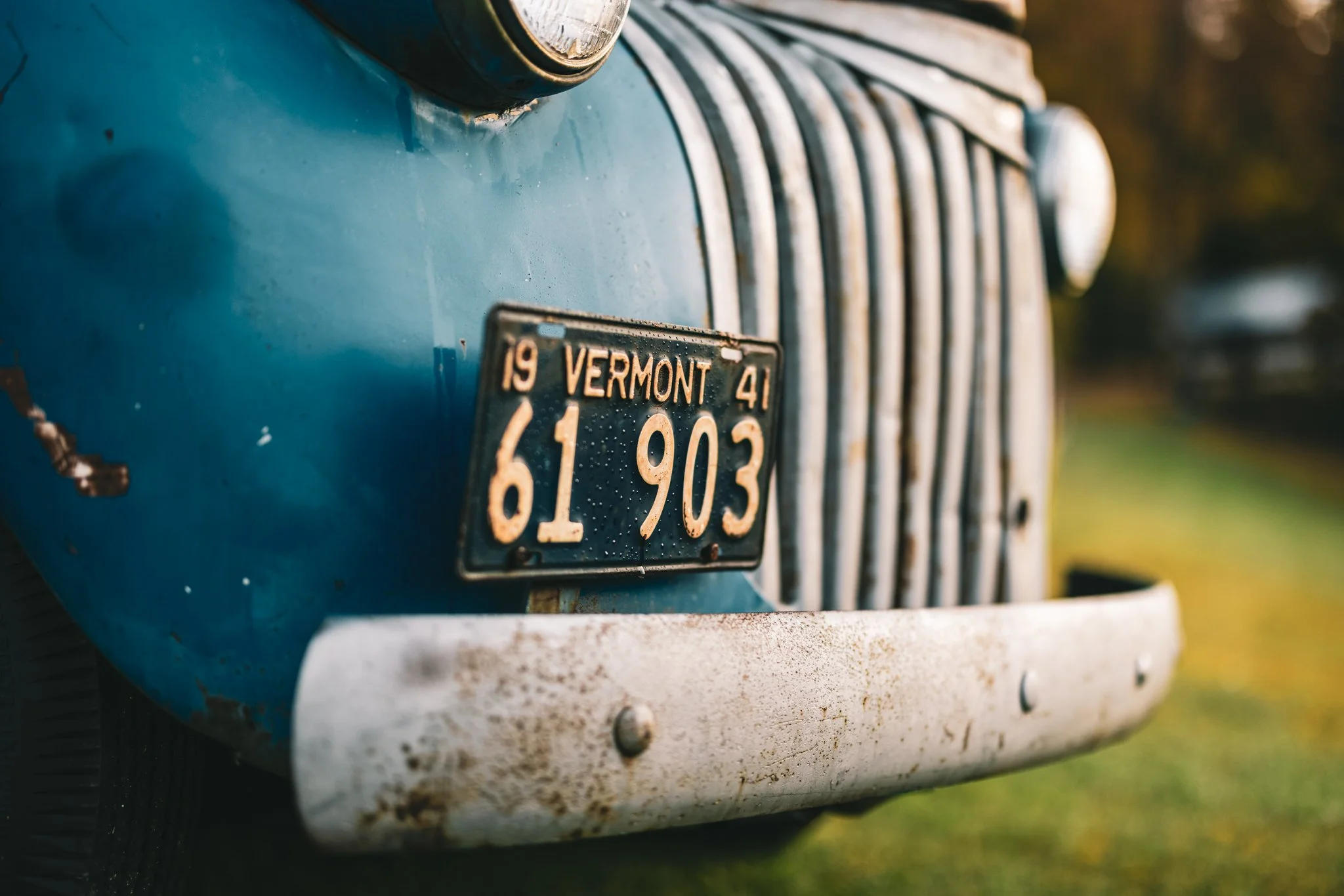 Close-up of the front of an old blue Chevy Pickup with a Vermont license plate, showing signs of age and rust.