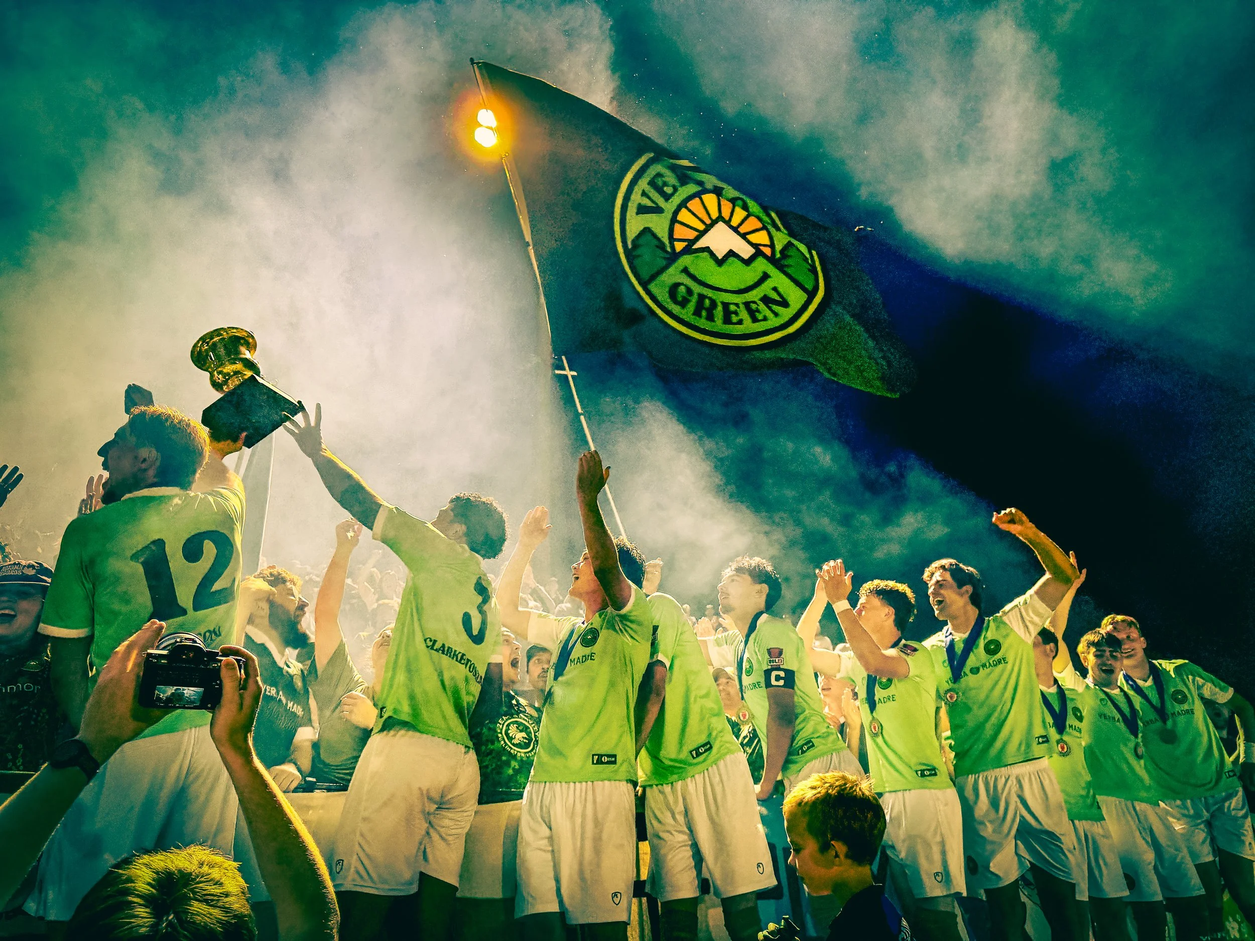 Group of sports players wearing yellow jerseys celebrating, with one holding a flag with a logo of a sun, mountains, and the word 'Green.' Some players are wearing medals, and there are audience members and photographers capturing the moment.