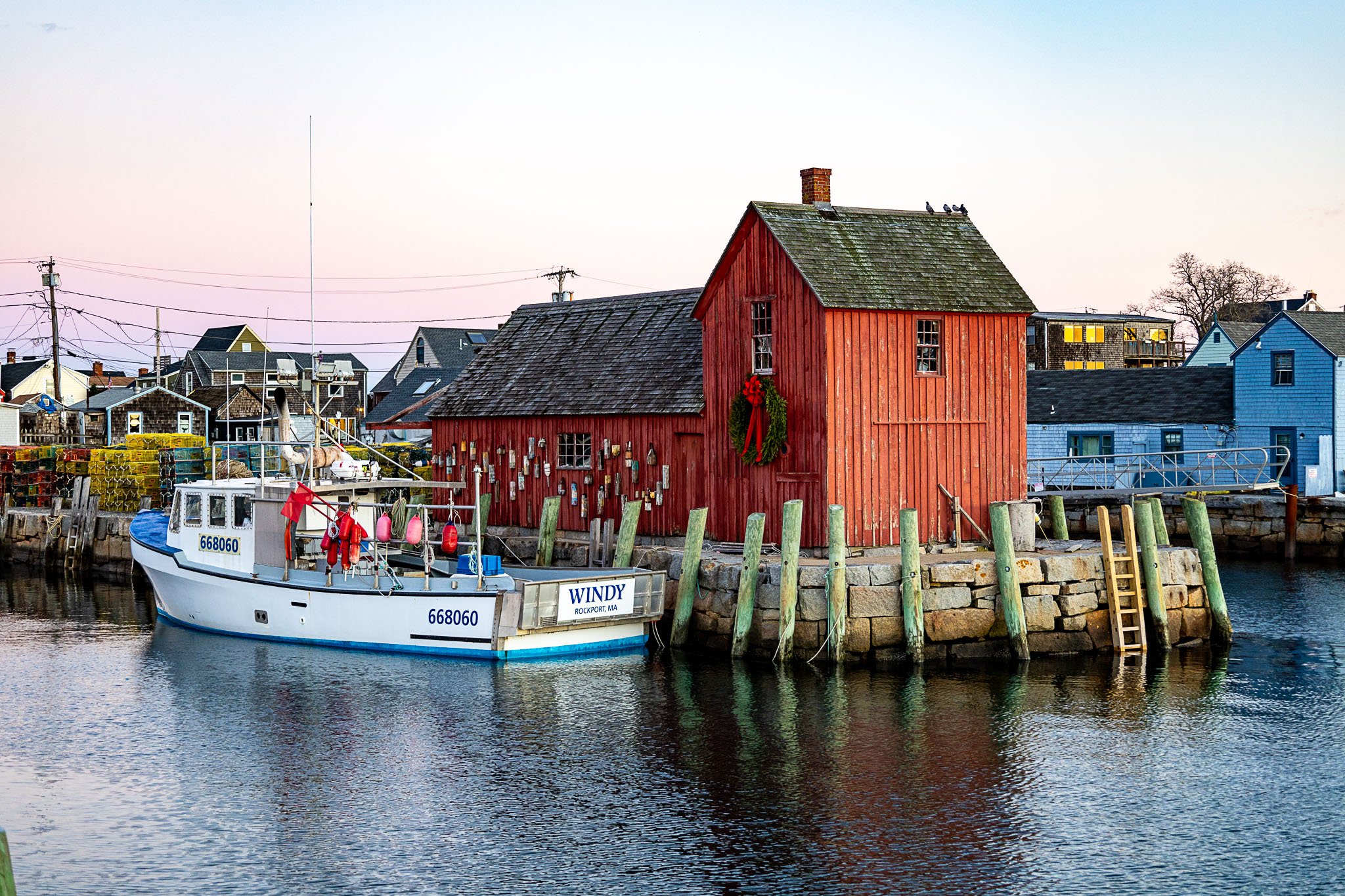 A small white boat named 'Windy' docked at a harbor, with a red wooden building decorated with Christmas wreath and stockings nearby. Multiple houses and buildings are visible in the background.