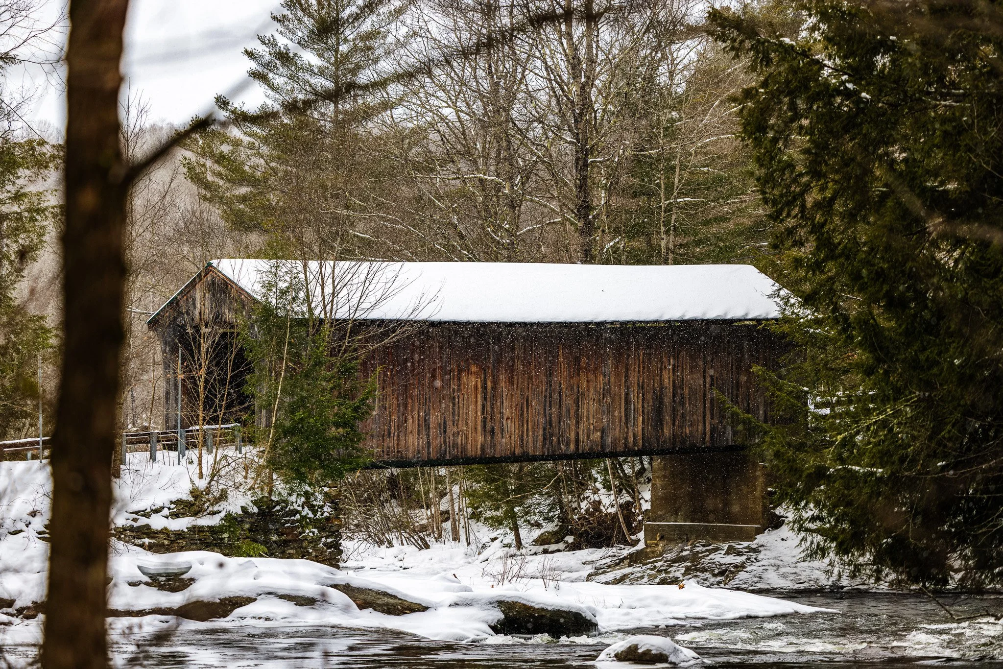 A covered wooden bridge over a stream in a snowy, wooded landscape, with trees and snow-covered ground surrounding it.