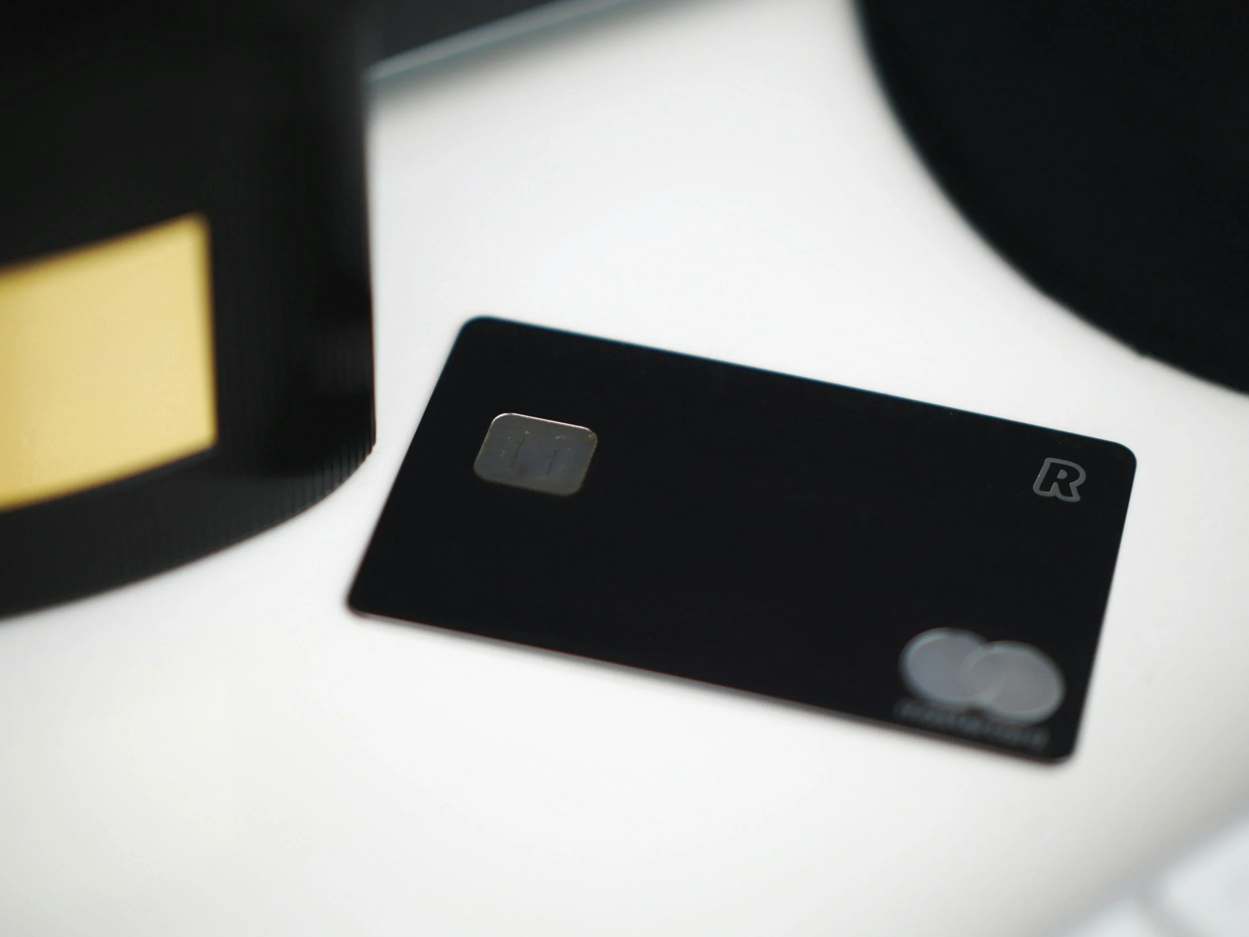 A black credit card with an R logo on the top right corner and a Mastercard logo on the bottom right corner, placed on a white surface, with part of a black object with yellow behind it visible in the top left corner.