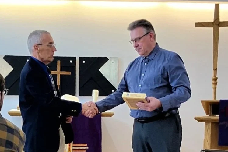 Two men shaking hands inside a church with a cross on the wall behind them.