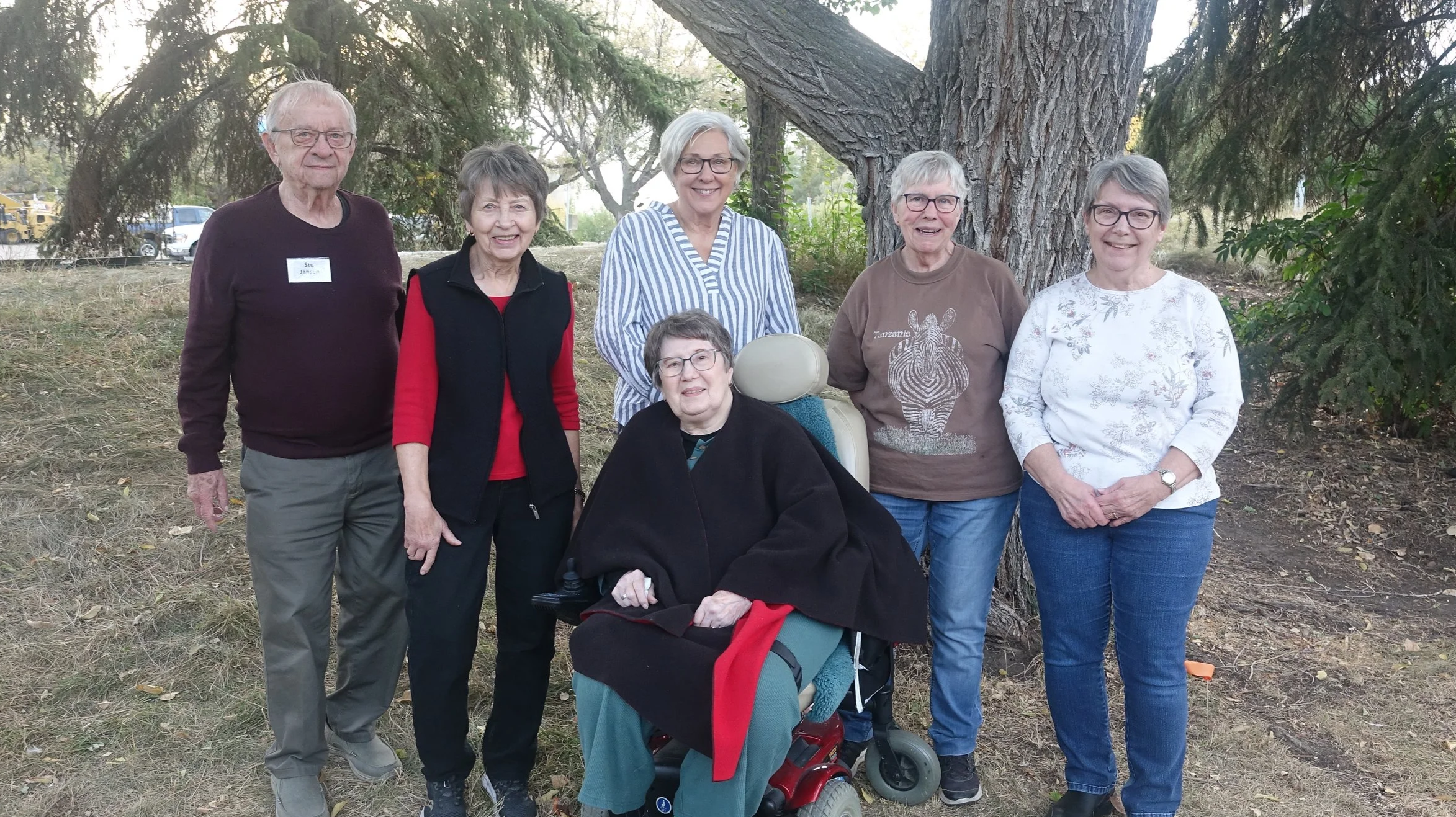 A group of seven seniors, six women and one man, standing outdoors near a large tree, with one woman seated in a motorized wheelchair. They are smiling and posing for the photo in a park-like setting with trees and parked cars in the background.