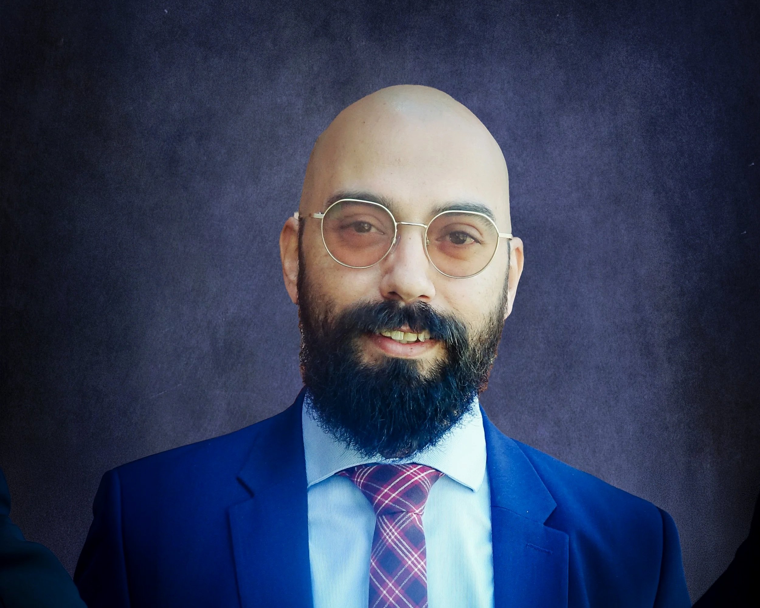Professional headshot of a bald man with a beard, wearing glasses, a dark blazer, pink shirt, and red plaid tie, smiling against a dark blue background.