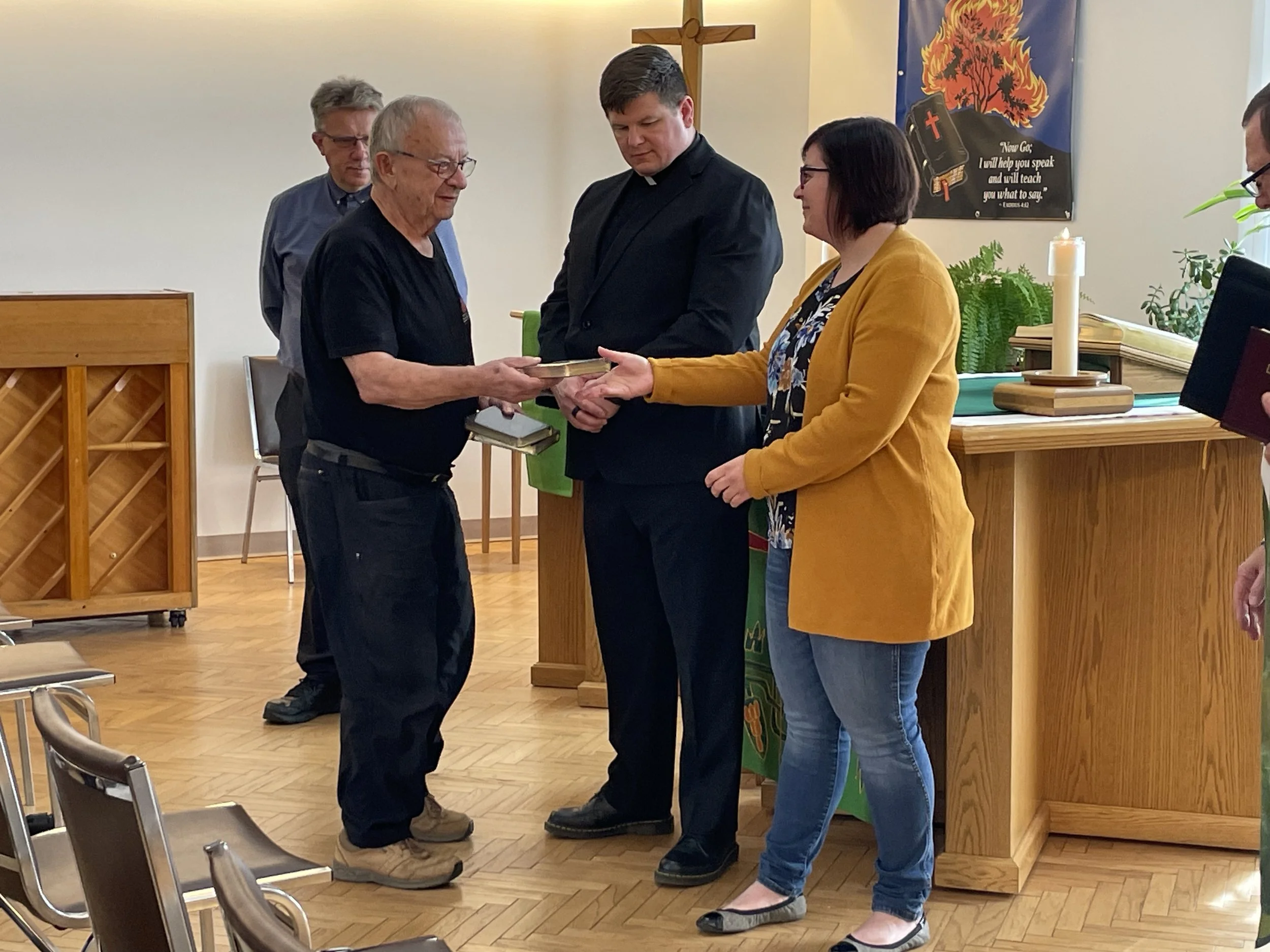 A man in a black t-shirt is receiving an award or certificate from a woman in a yellow blazer, while a man in a clerical outfit and another man in glasses stand nearby during a ceremony in a church setting.