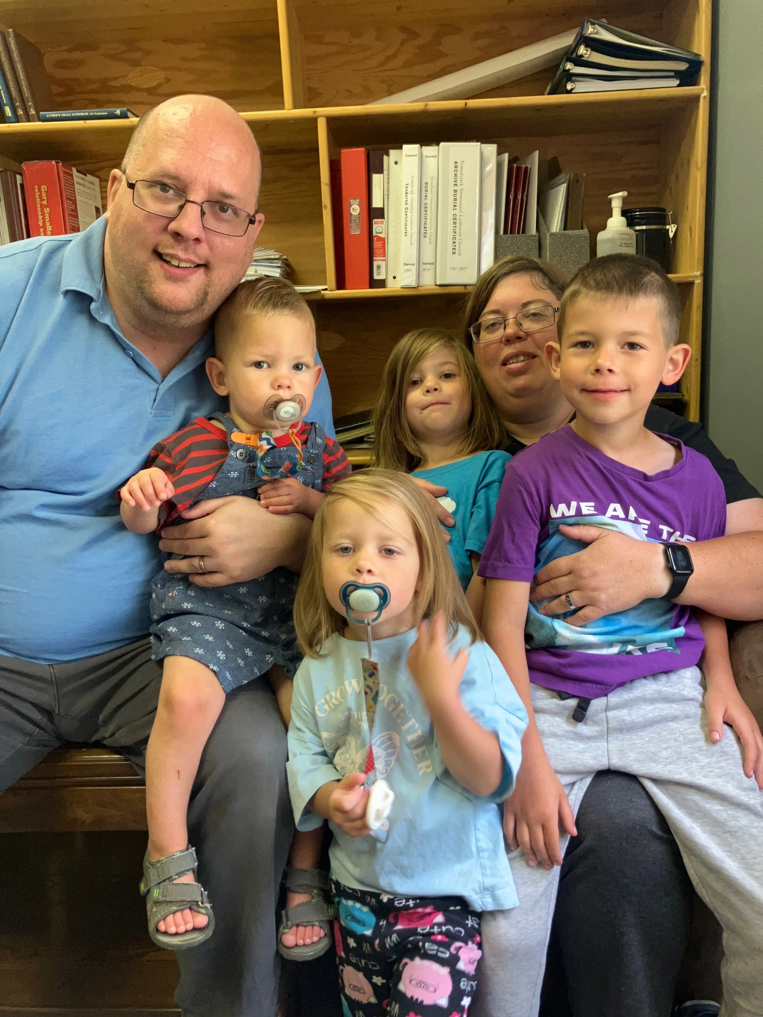 A family group photo indoors with three adults and four children, some with pacifiers, sitting and standing close together, in front of wooden bookshelves with books, binders, and medical supplies.