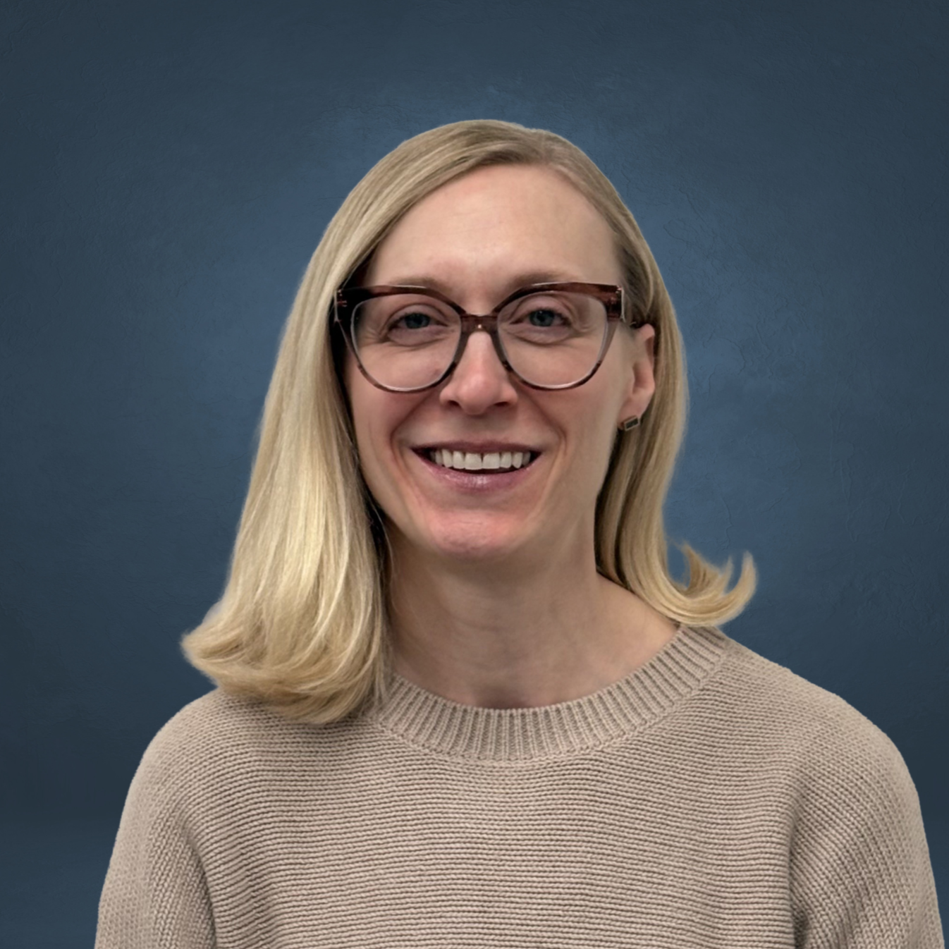 A woman with blonde hair and glasses smiling, wearing a beige sweater, standing against a dark blue background.