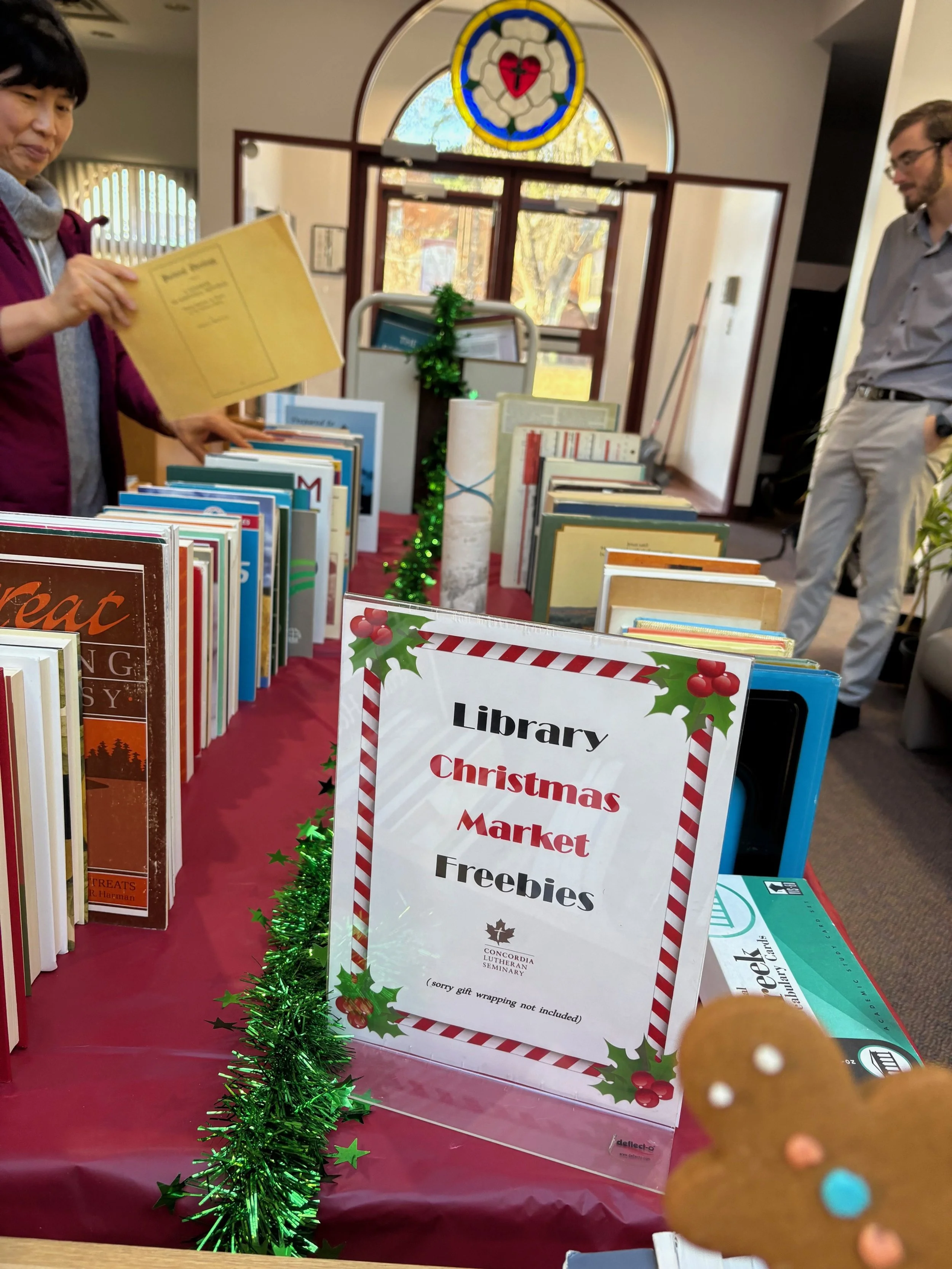 Table with Christmas-themed sign promoting free books at the library's Christmas market. Decorated with green tinsel and holly, surrounded by books. People are browsing in the background inside a church or community center with stained glass window.