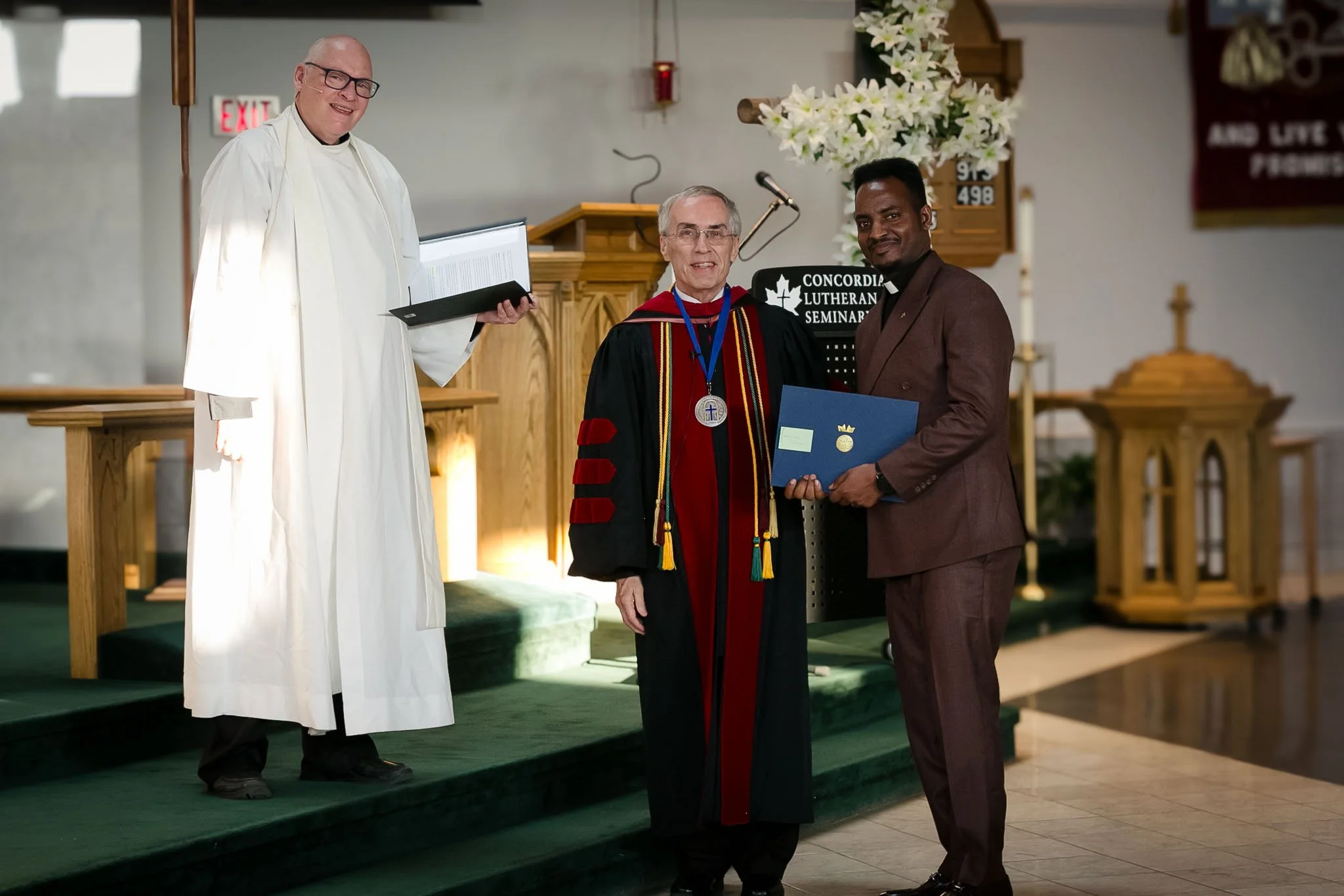 Three men standing on a stage inside a church or seminar room. One in religious robes, one in academic regalia, and one in a suit, holding a certificate and smiling for the photo.
