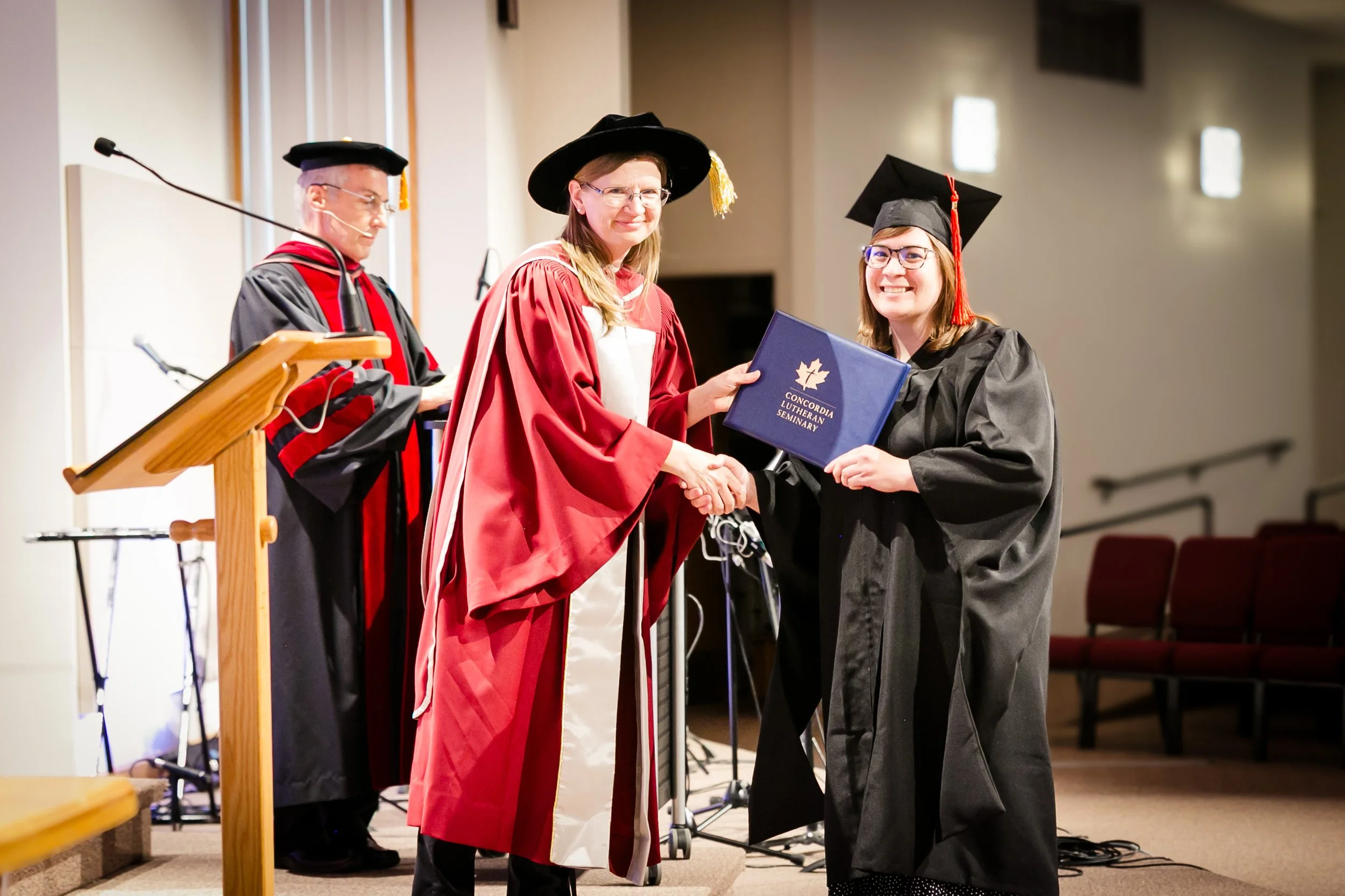 A graduation ceremony where a woman in a red academic gown and cap is shaking hands with a graduate in a black gown and cap, holding a diploma from Concordia Lutheran Seminary. An official in a gown and cap stands nearby.