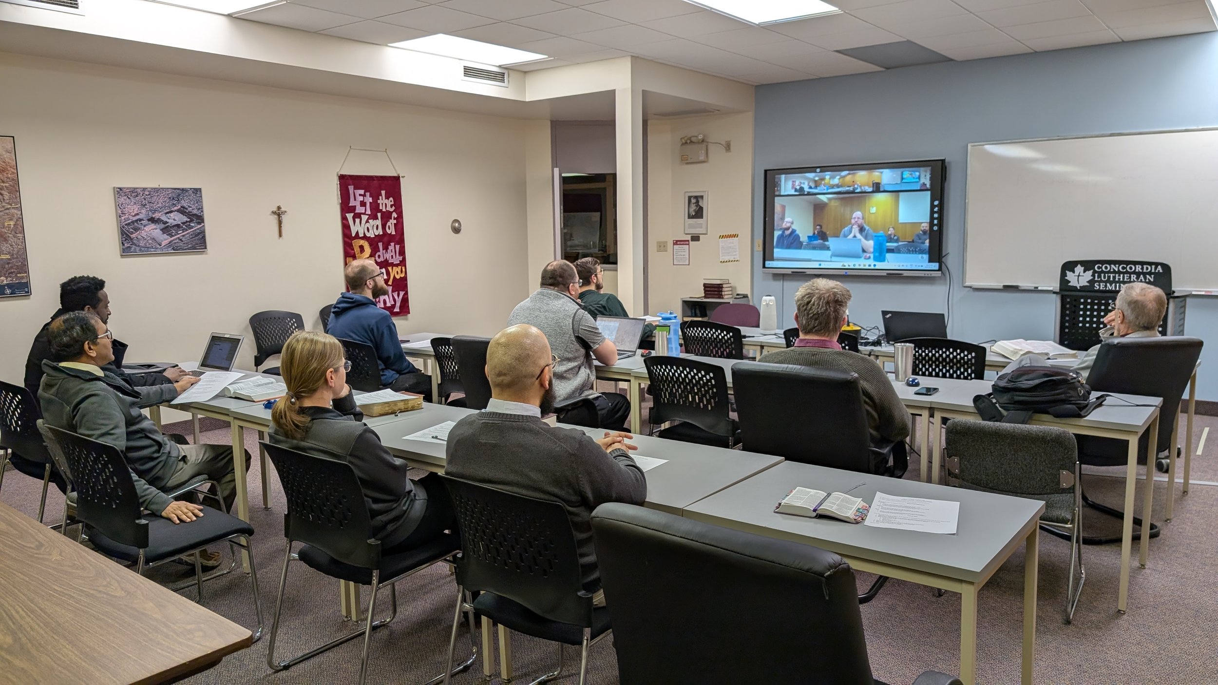 A classroom with students sitting at tables watching a video call on a large wall-mounted screen. The room has books, papers, and laptops on the tables, with a whiteboard and a banner on the wall.