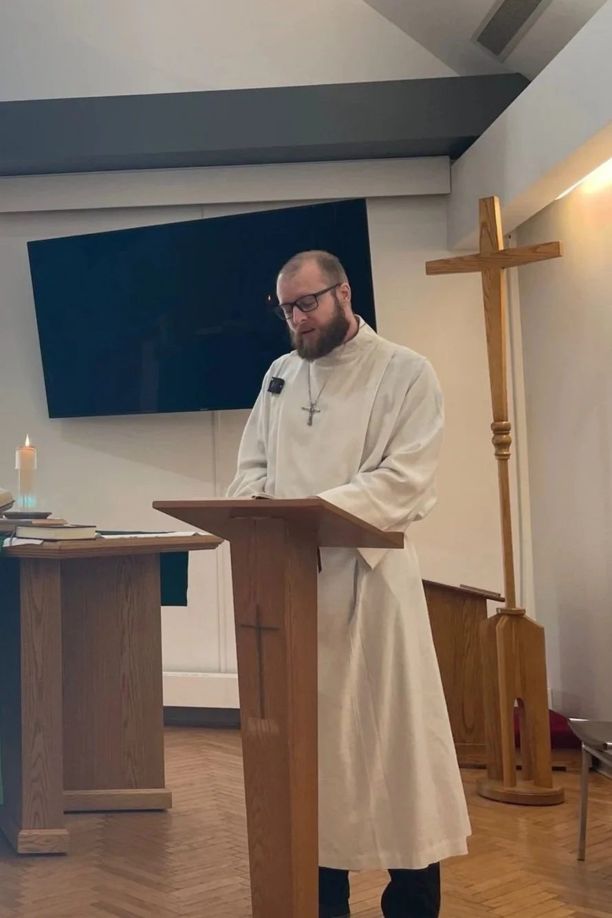 A man dressed in a white religious robe with a cross necklace and glasses is standing at a wooden lectern with a cross symbol, inside a church or chapel. Behind him, there is a large flat-screen TV mounted on the wall and a wooden cross standing upright. A small lit candle is on a table nearby.