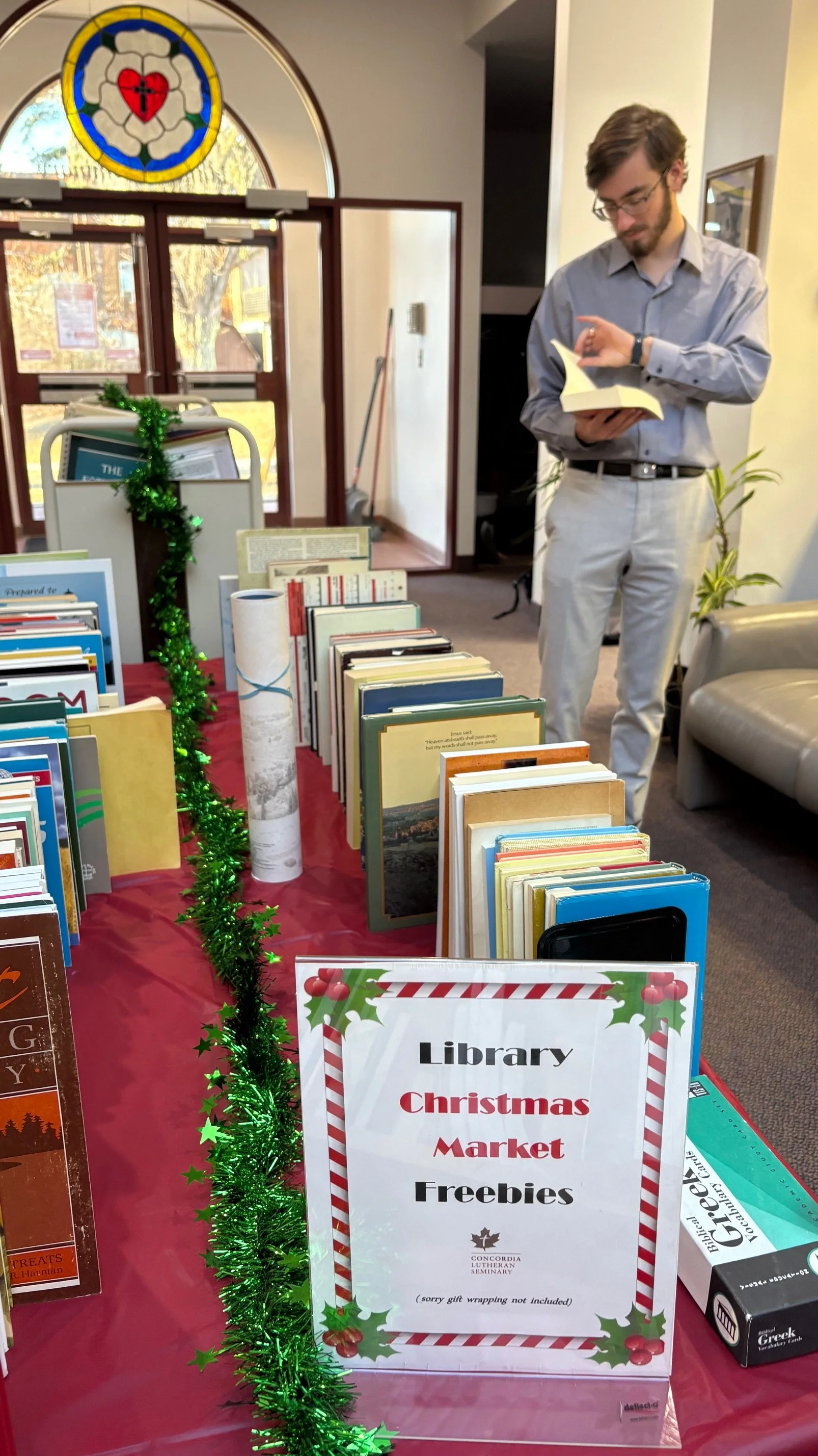 A man standing next to a table filled with Christmas-themed books and freebie signs at a library. Decorated with green tinsel, the event is titled 'Library Christmas Market Freebies.'