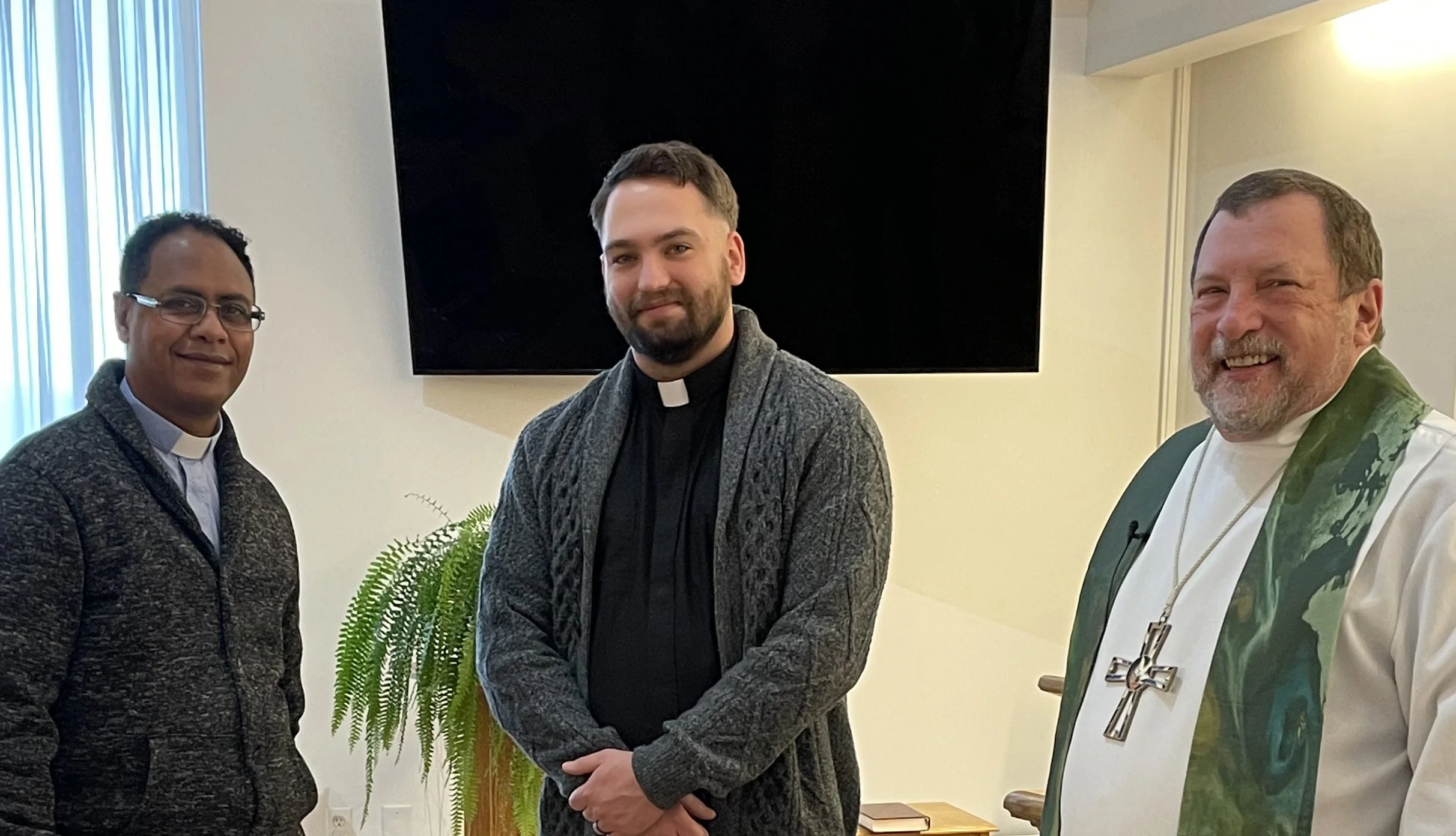 Three men, two in religious clerical attire, standing indoors in front of a wall with a large black screen and a potted fern plant, smiling at the camera.