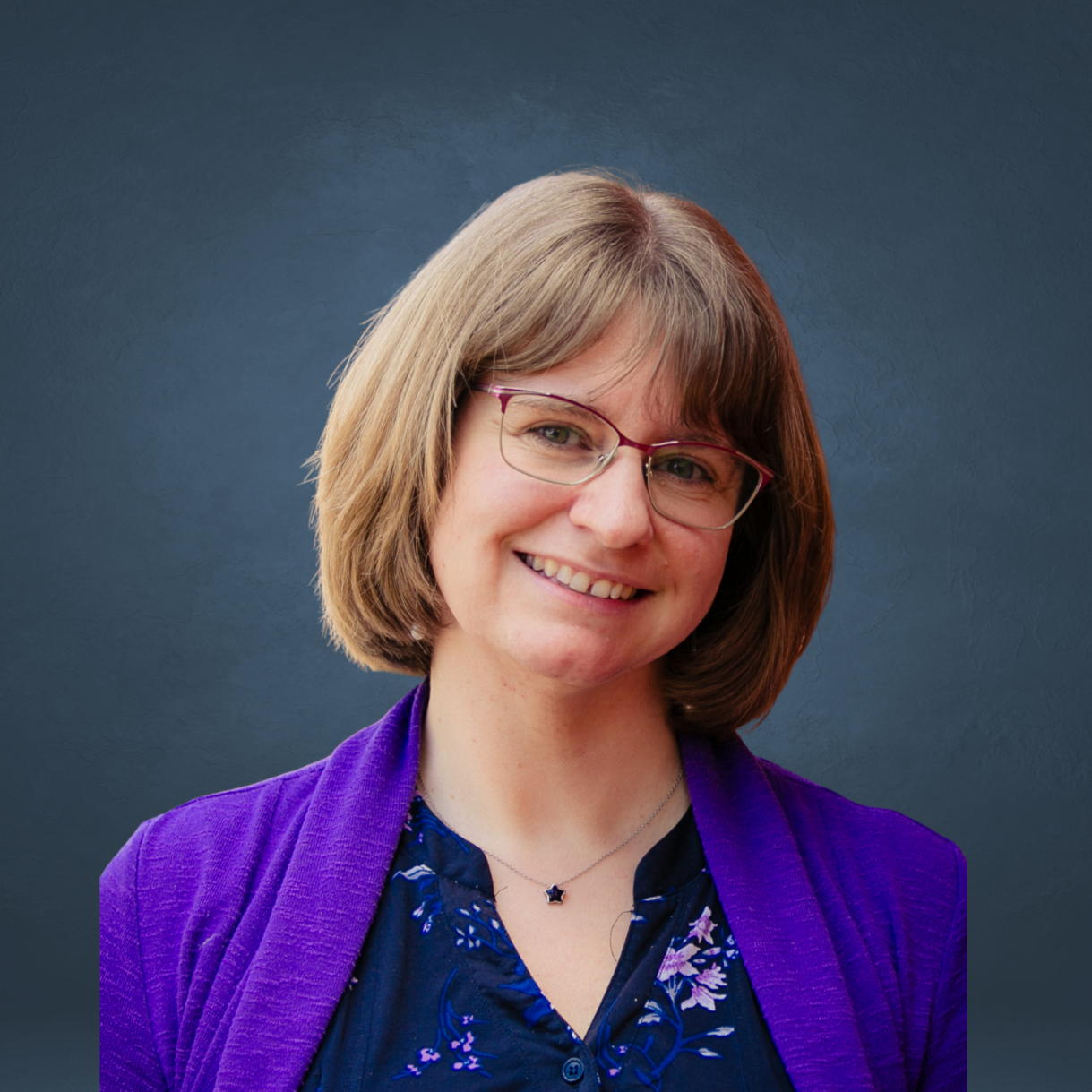 Smiling woman with shoulder-length light brown hair, glasses, wearing a purple blazer and a blue floral blouse against a dark blue background.
