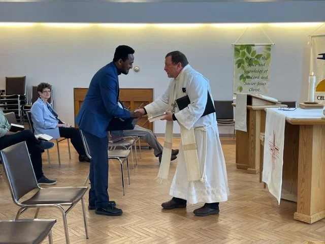 A priest and a man in a blue suit shaking hands in a church, with seated attendees in the background.