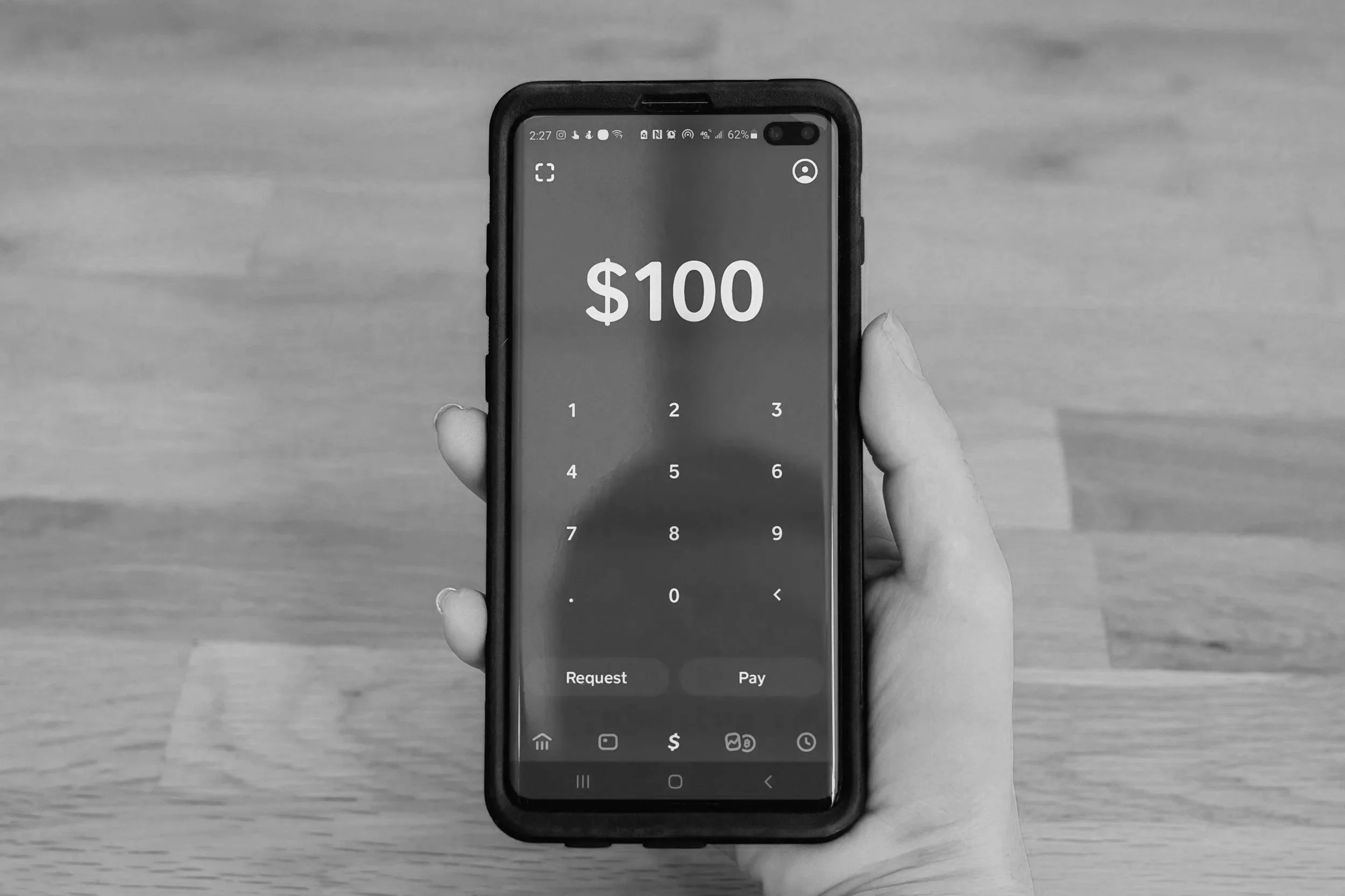 A person holding a smartphone displaying a mobile payment app with a $100 transfer screen on a wooden surface background.