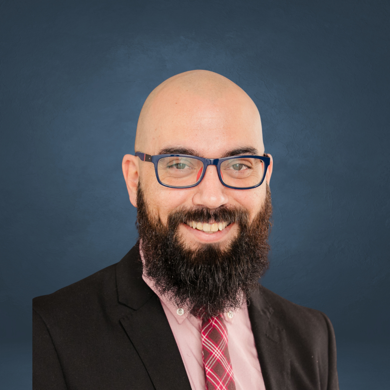 Professional headshot of a bald man with a beard, wearing glasses, a dark blazer, pink shirt, and red plaid tie, smiling against a dark blue background.