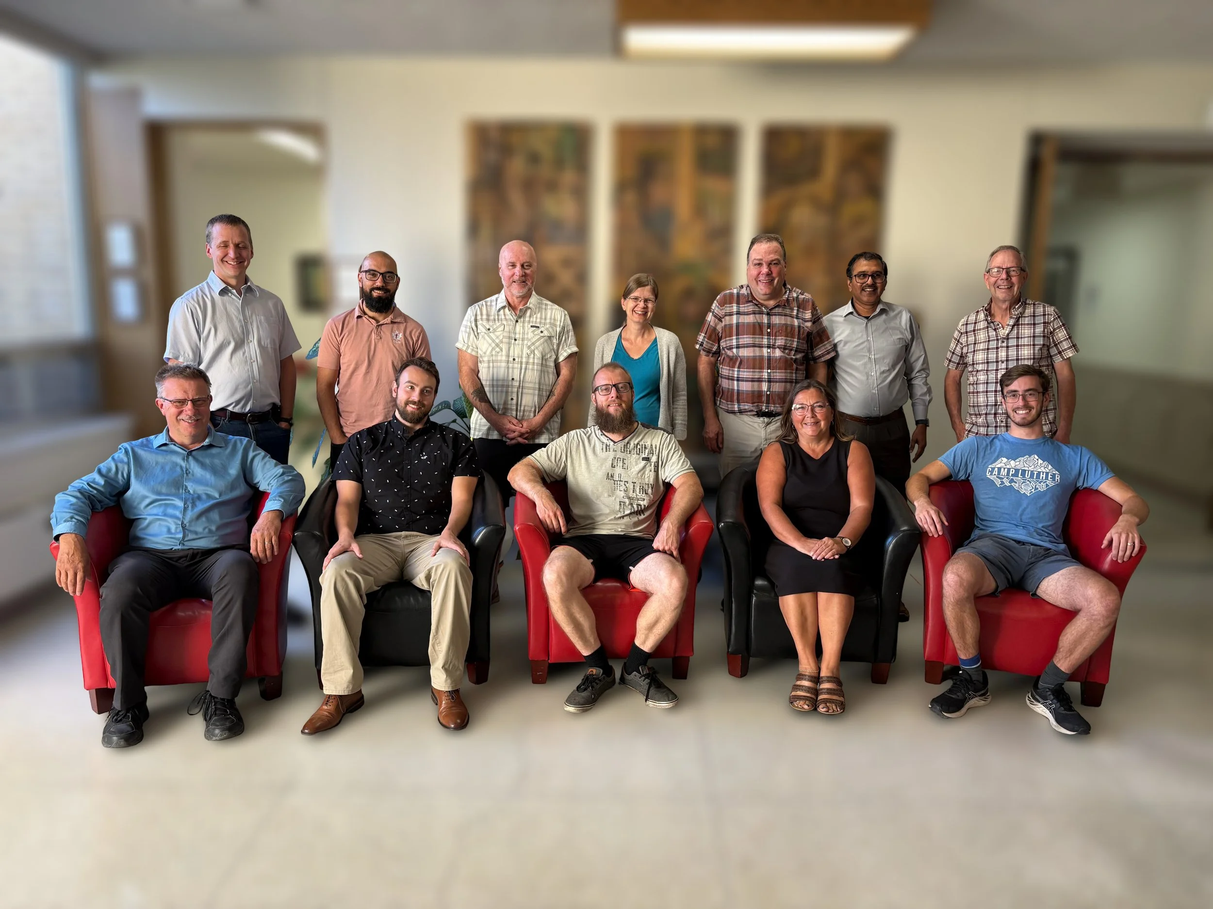 Group of 13 diverse adults posing for a photo in an indoor setting, some sitting in chairs and others standing behind them.