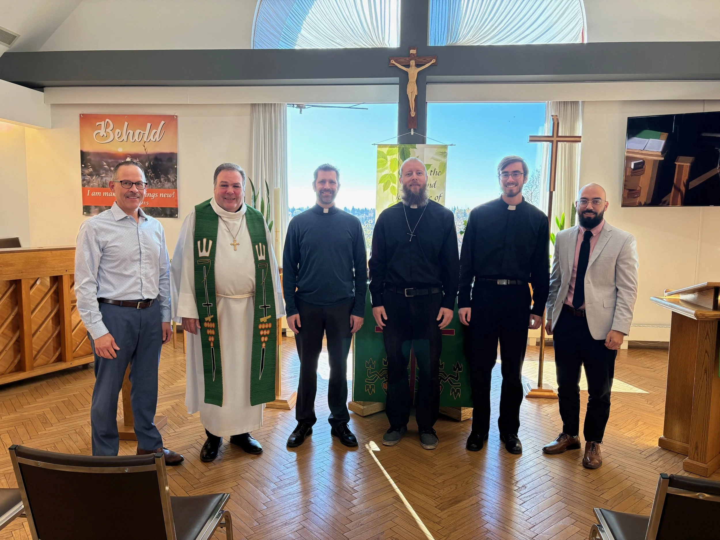 Six men standing inside a church, with a large wooden crucifix above them, two of whom are priests wearing clerical robes and collars, and the others in formal attire, posing in front of a window with sunlight coming through.