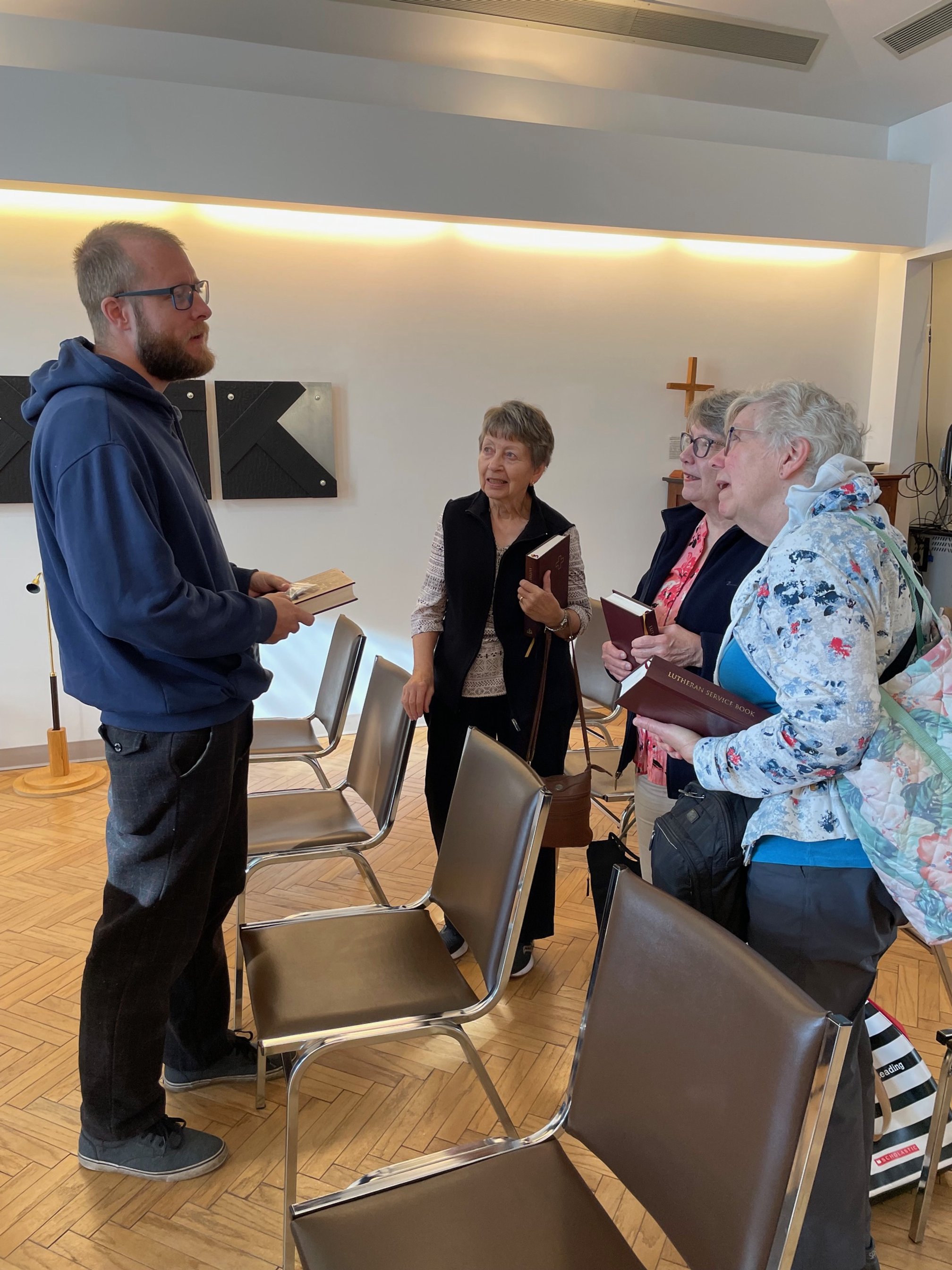 Four older women and one younger man engaged in conversation inside a chapel or church, with hymn books in their hands, a cross on the wall, and chairs arranged for a gathering.