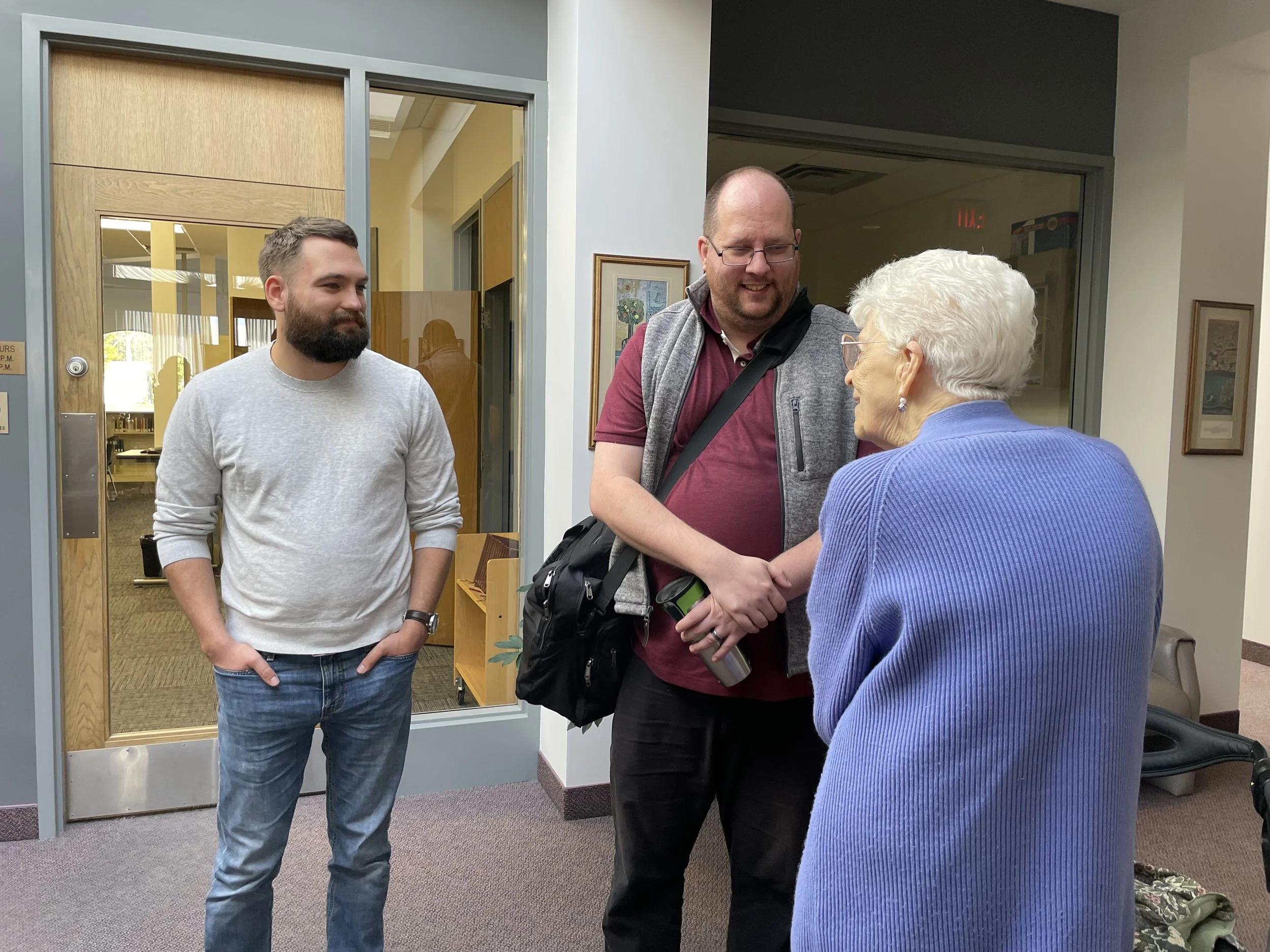Two men shaking hands with an elderly woman in an indoor setting, with one man standing slightly behind and smiling at the interaction.