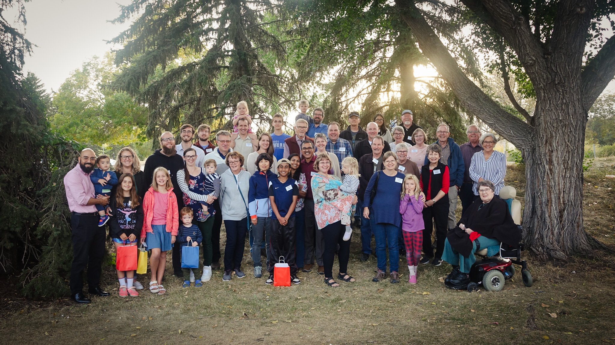 A large group of people, including children, adults, and elderly, gathering outdoors under trees for a group photo. The sunlight is setting behind the trees, creating a warm glow.