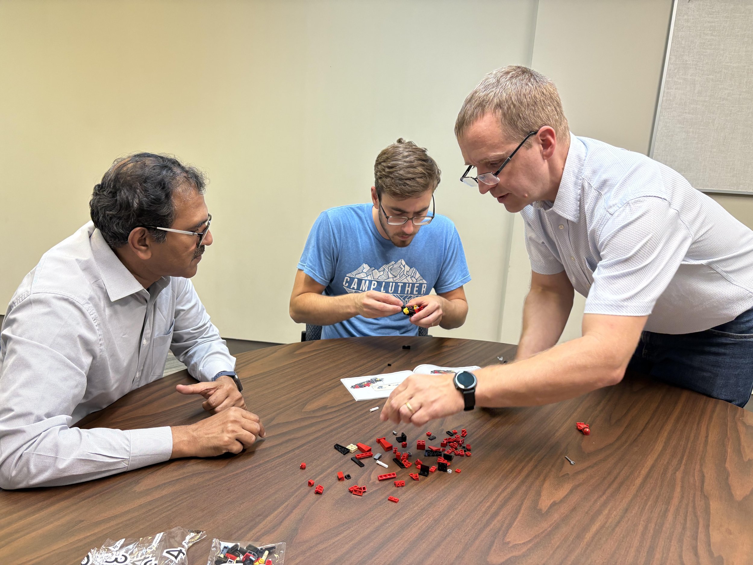 Three men are sitting at a wooden table assembling a LEGO set. The man on the left is wearing glasses and a light gray shirt, the man in the middle is wearing glasses and a blue T-shirt, and the man on the right is wearing glasses and a white shirt. 