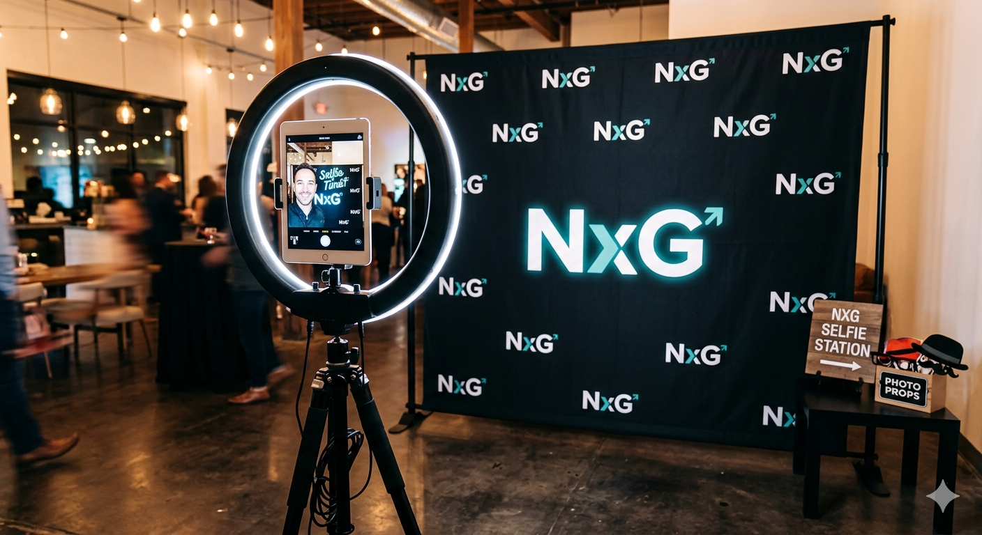 Photo booth setup with a ring light and a camera on a tripod in front of a step-and-repeat backdrop with 'NxG' logos, featuring an 'NXG Selfie Station' sign and props on a table.