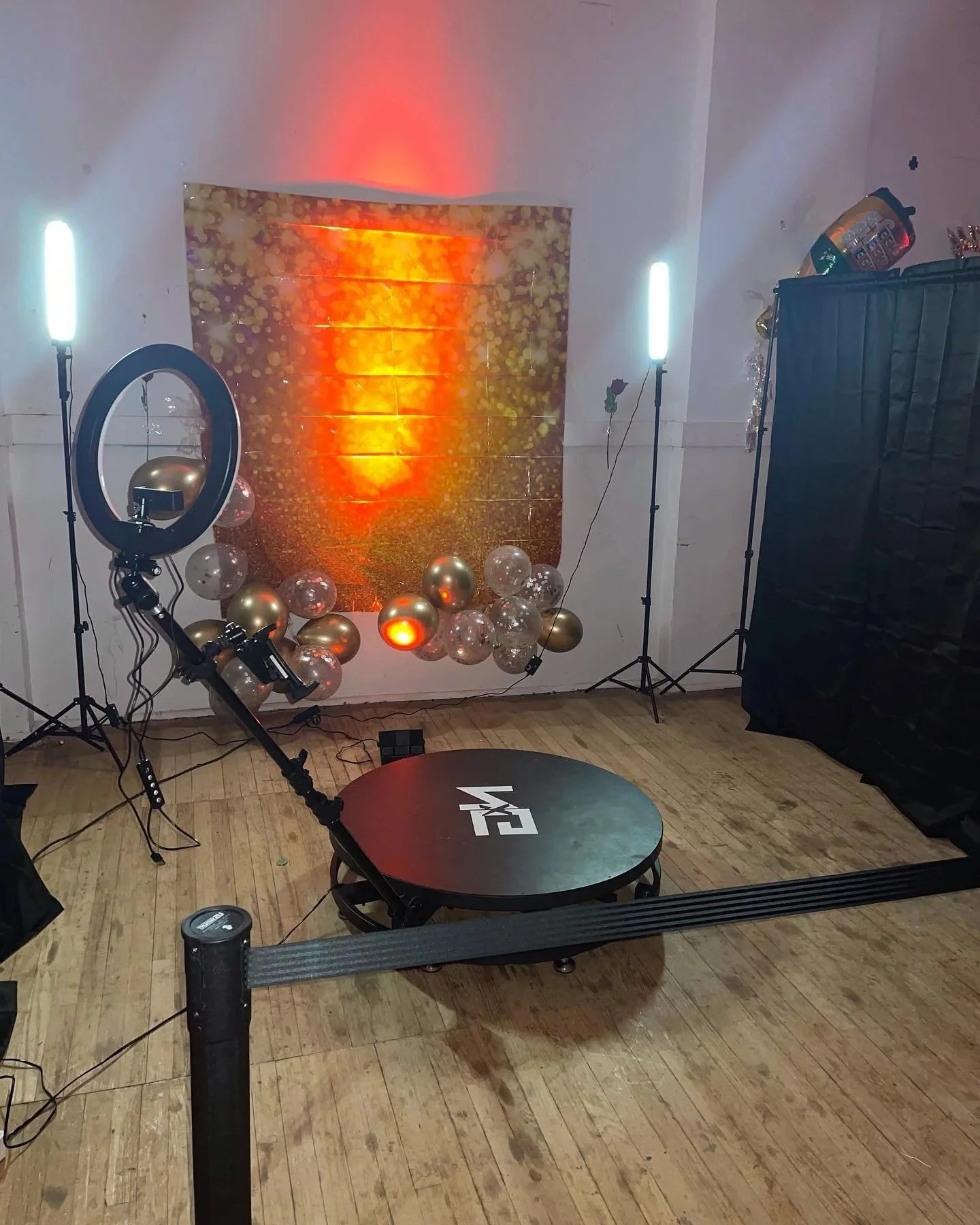 Photo of a photo booth setup with a ring light, balloon decorations, a backdrop, and professional lighting equipment on a wooden floor.