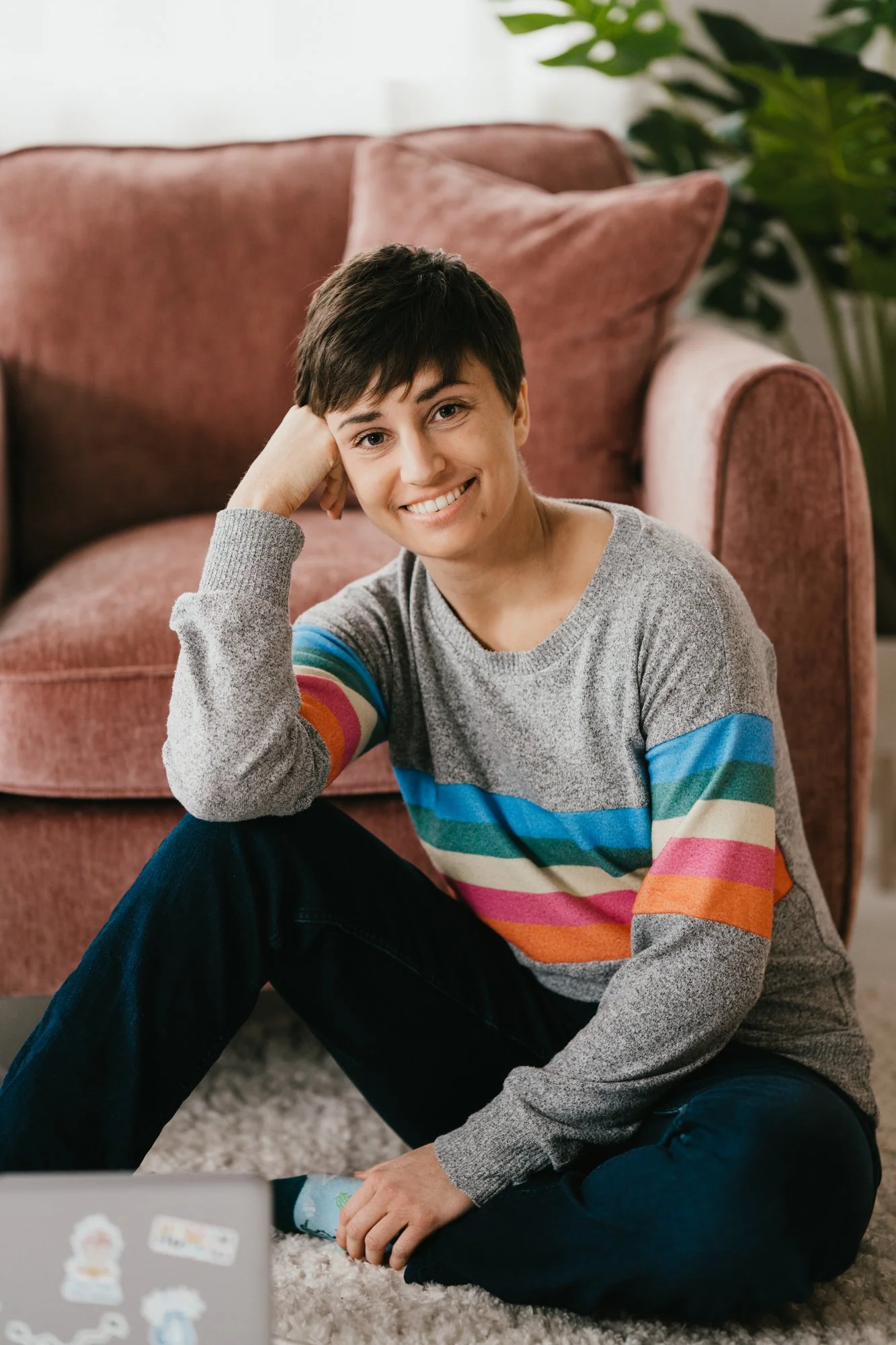 Jen, therapist, sitting on the floor, in front of a pink chair, one knee up, resting her elbow on her knee, and her face on her hand. Smiling at the camera wearing a rainbow jumper.
