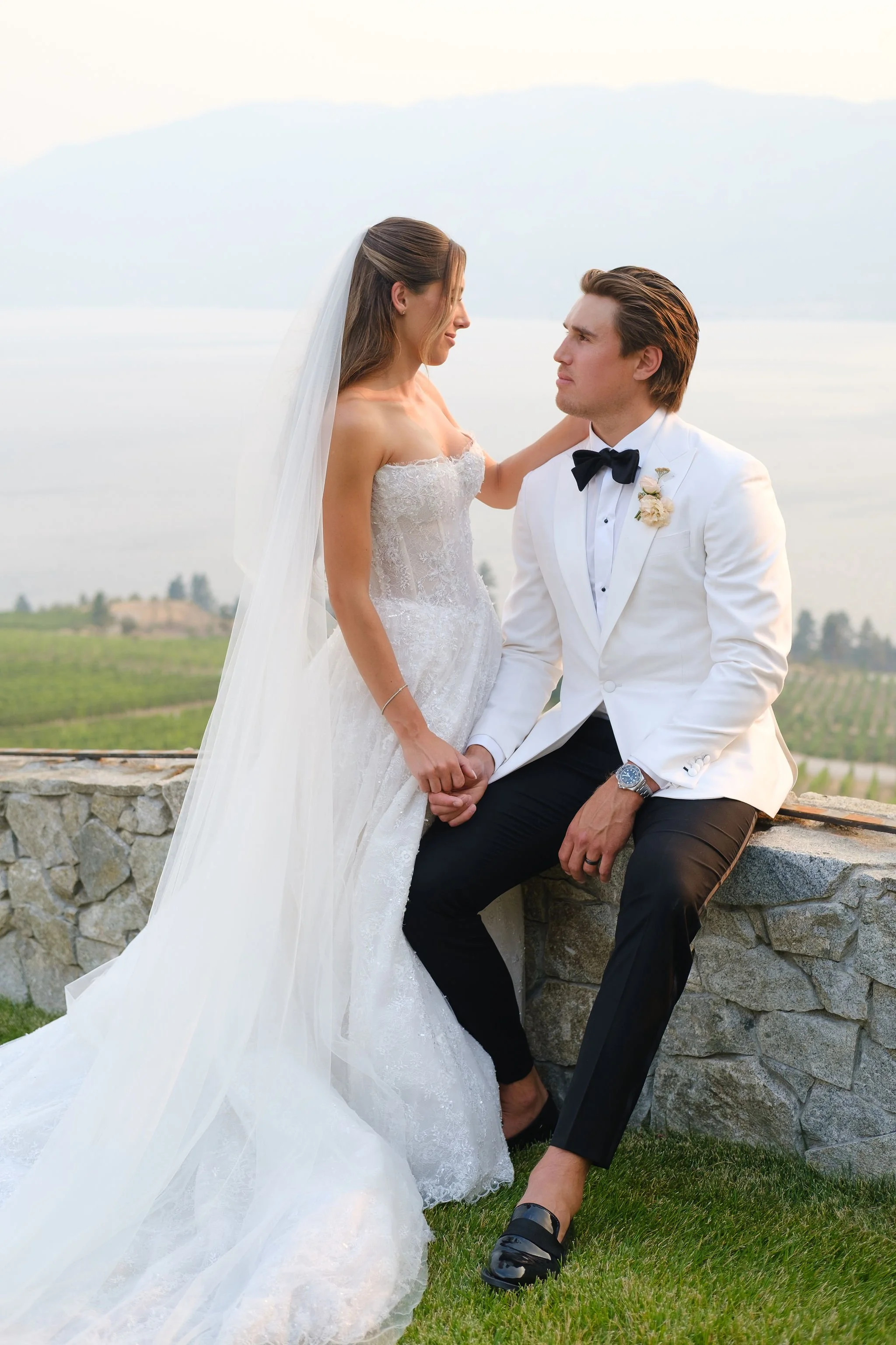 A bride and groom pose outdoors with a scenic background, the bride in a white wedding gown with a veil and the groom in a white tuxedo with a black bow tie and boutonniere, holding hands and looking at each other with serious expressions.