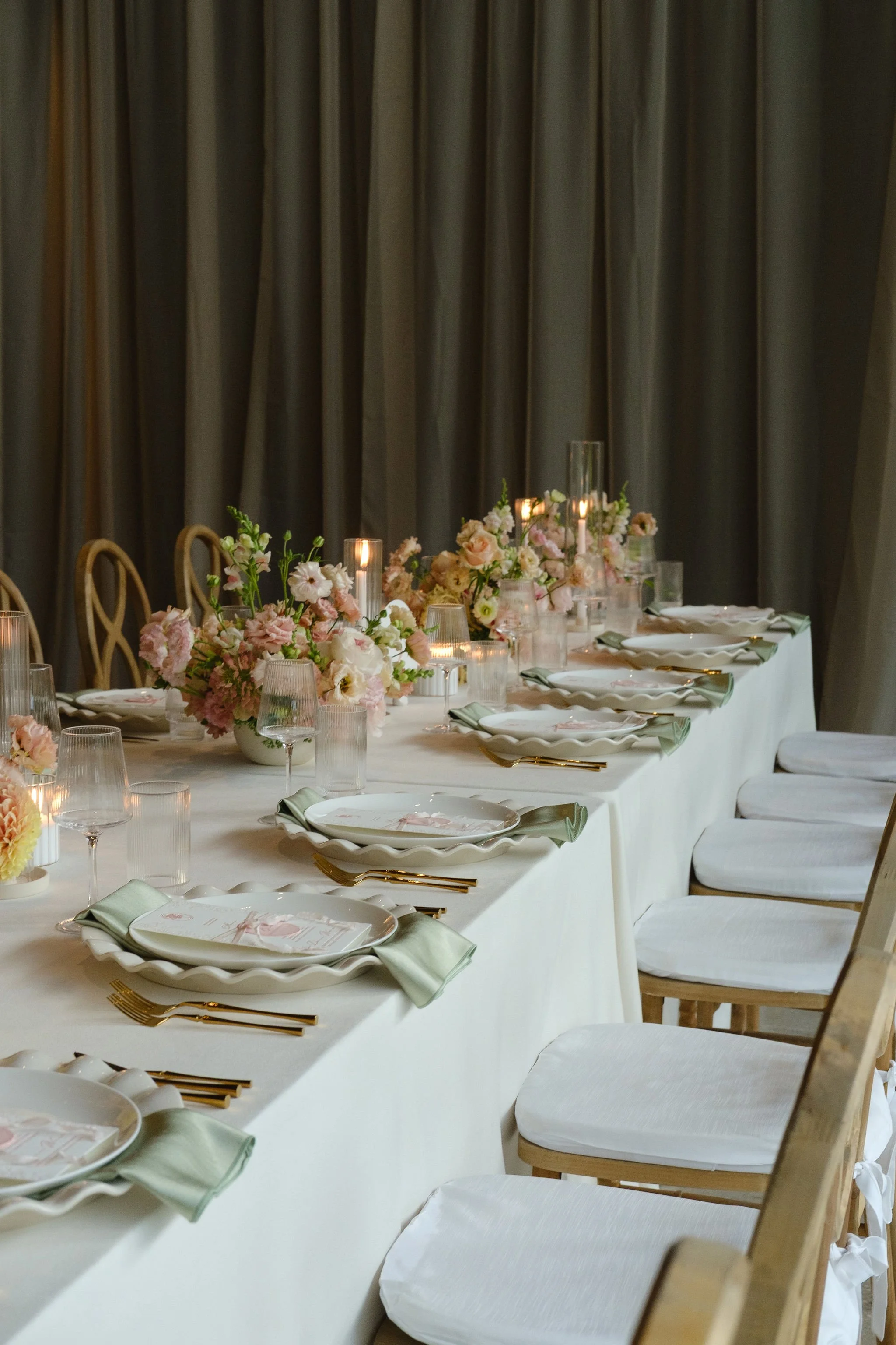A long dining table decorated with pink and white flowers, candles, and tableware, with chairs on one side and a dark curtain backdrop.