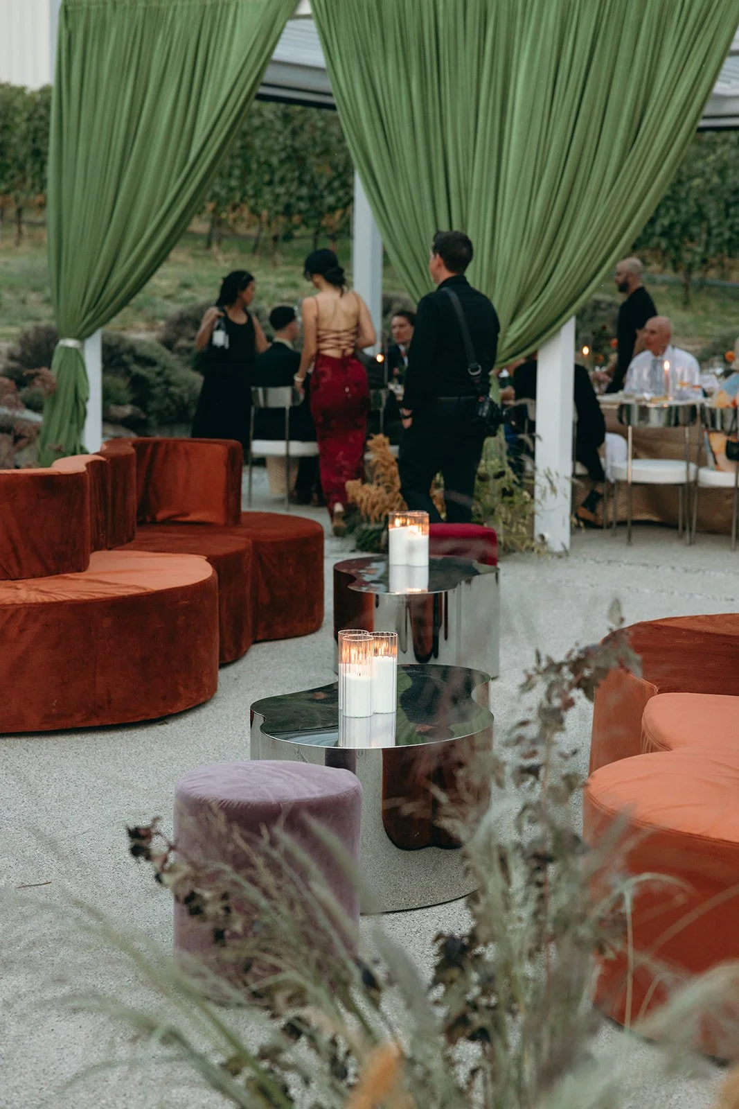 Outdoor event with people gathered, green drapes, modern tables with candles, velvet seating, and a vineyard in the background.