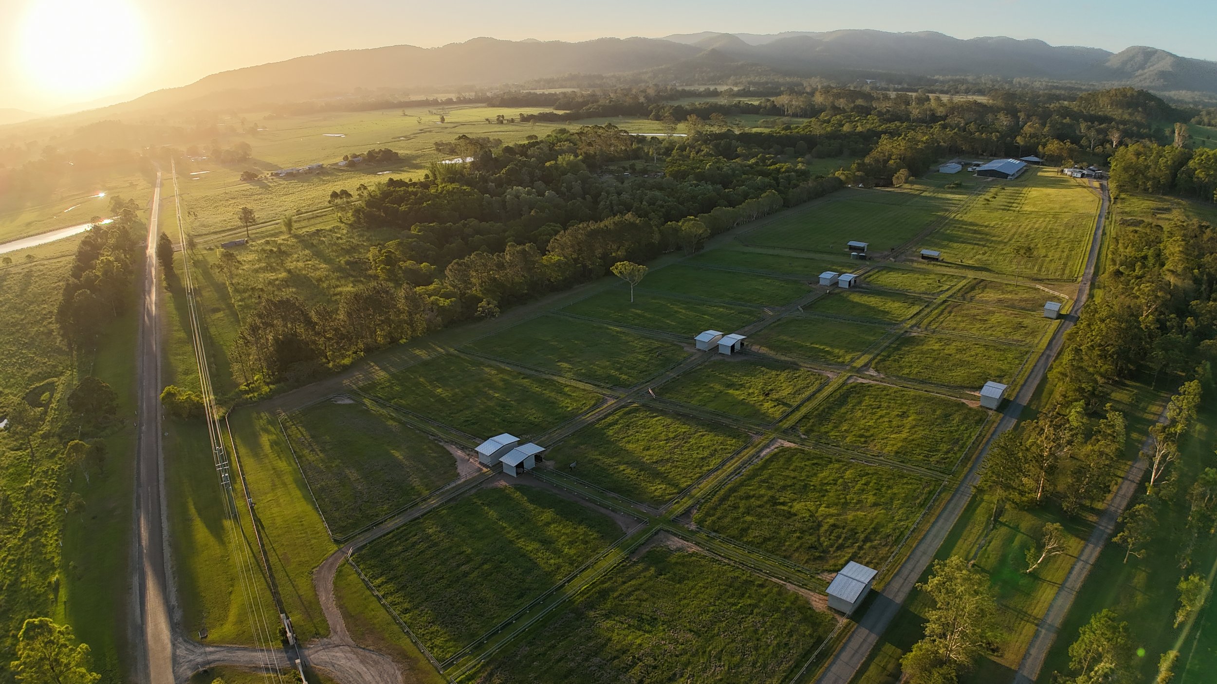 Aerial view of a rural farm at sunset, showing organized farmland, small structures, a paved road, and scenic mountains in the background.