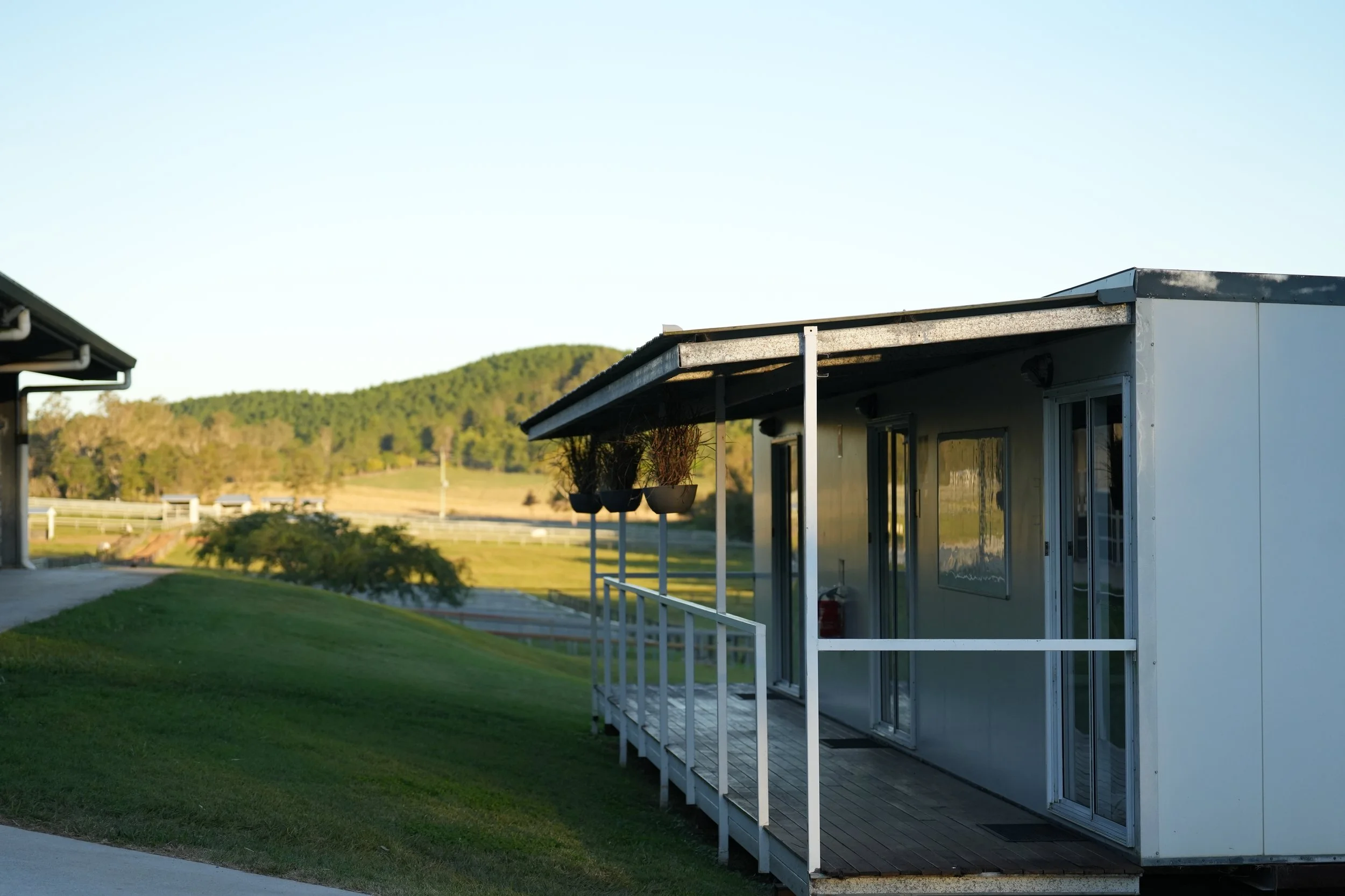 Small white house or trailer with a porch and hanging plants, on a grassy hillside with trees and hills in the background.