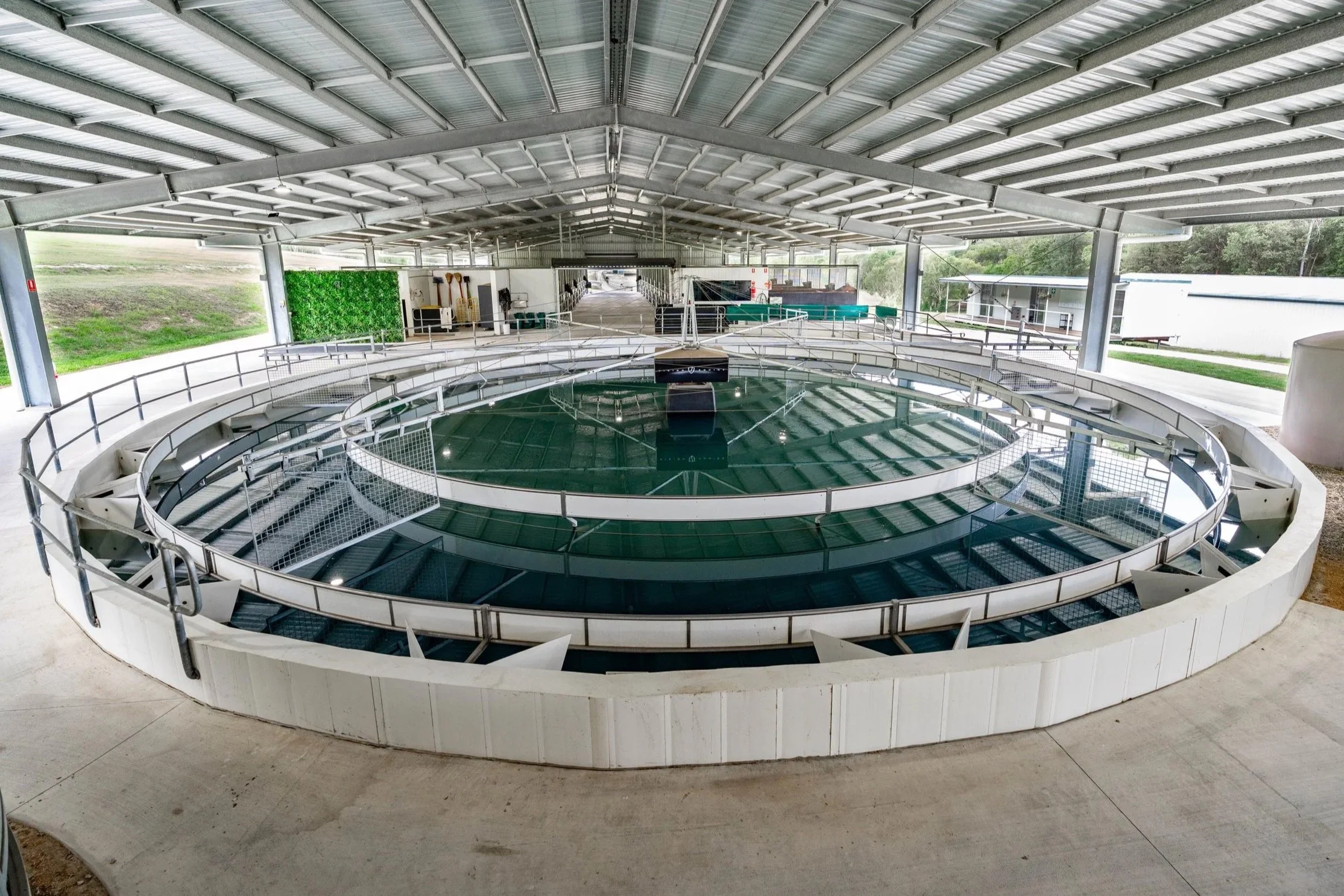 Indoor facility housing a circular fish tank with a reflective glass surface, surrounded by metal safety railings, with a roof overhead and outdoor greenery visible through openings.