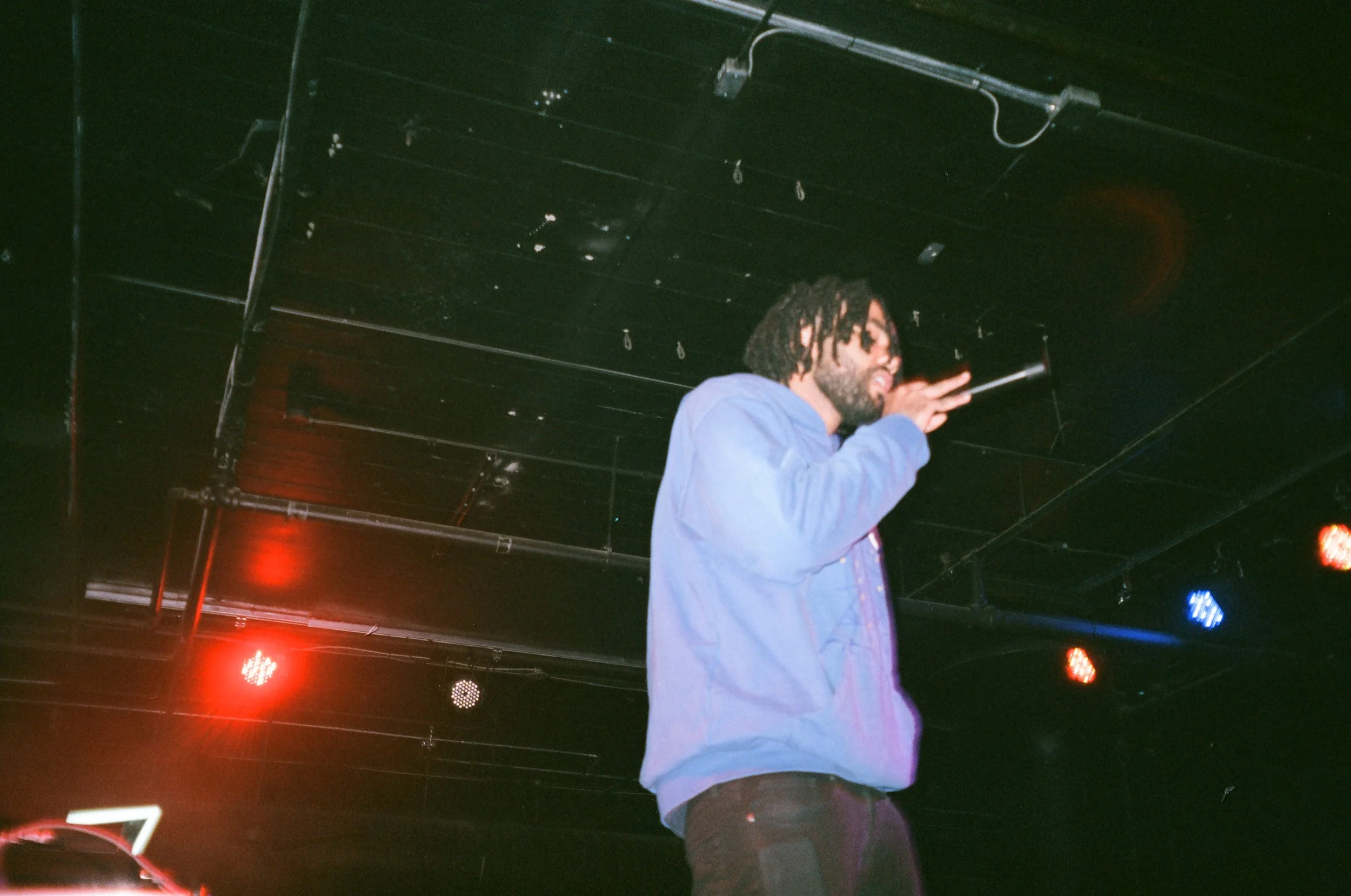 A man with dreadlocks and a beard, wearing a blue jacket, is holding a microphone and performing on stage with red and blue stage lights overhead.