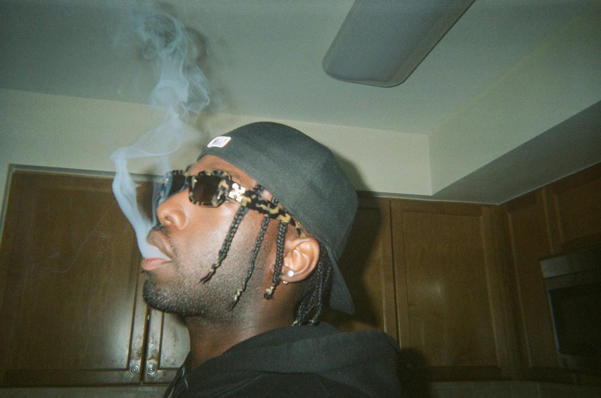 A man in a black cap and sunglasses is smoking, with smoke visible in the air, in a room with wooden cabinets.