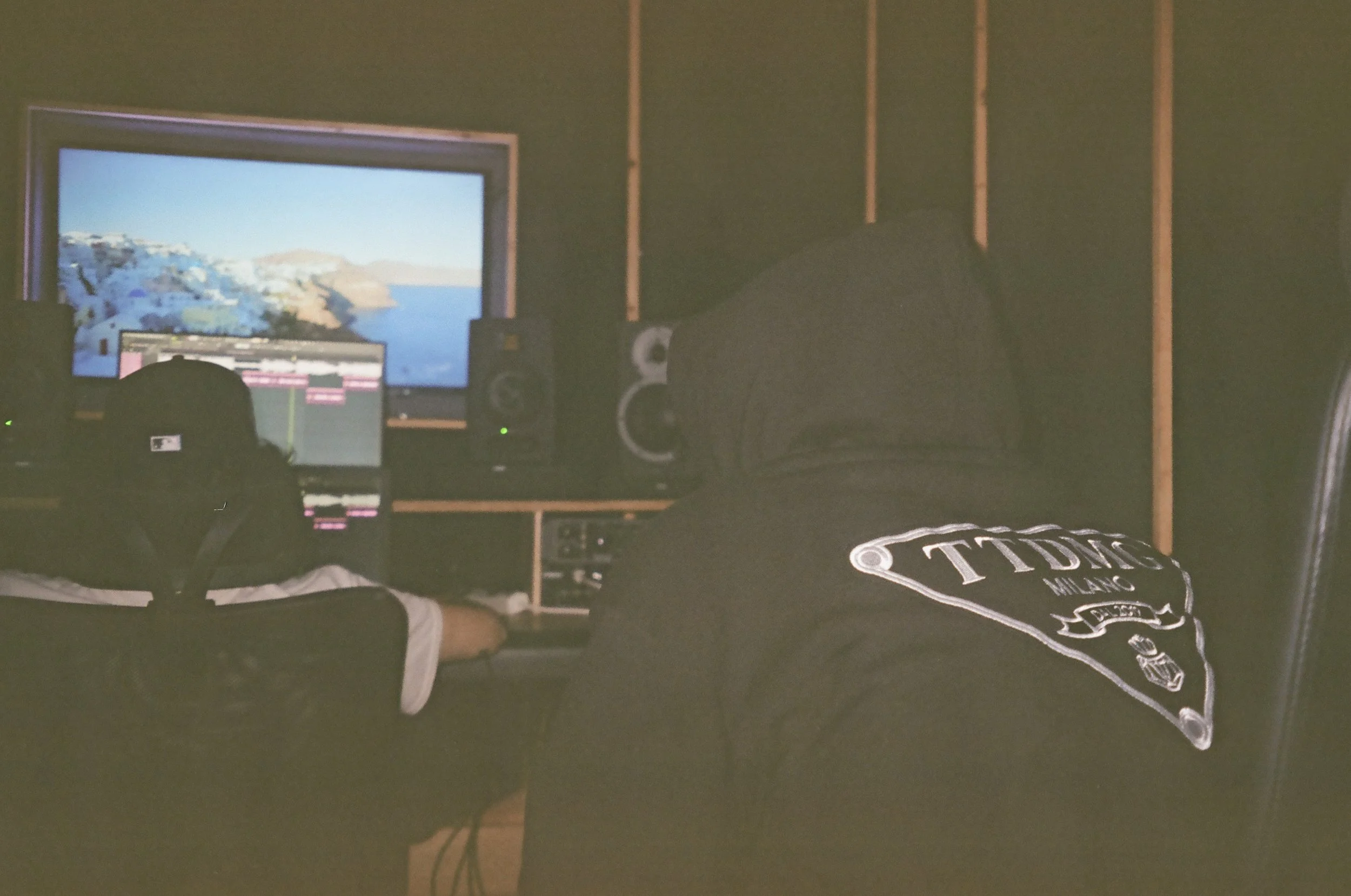 Two people in a recording studio working on music production. One person is sitting at a workstation with a computer and audio equipment, and the other is sitting in front of a large monitor displaying a scenic landscape.
