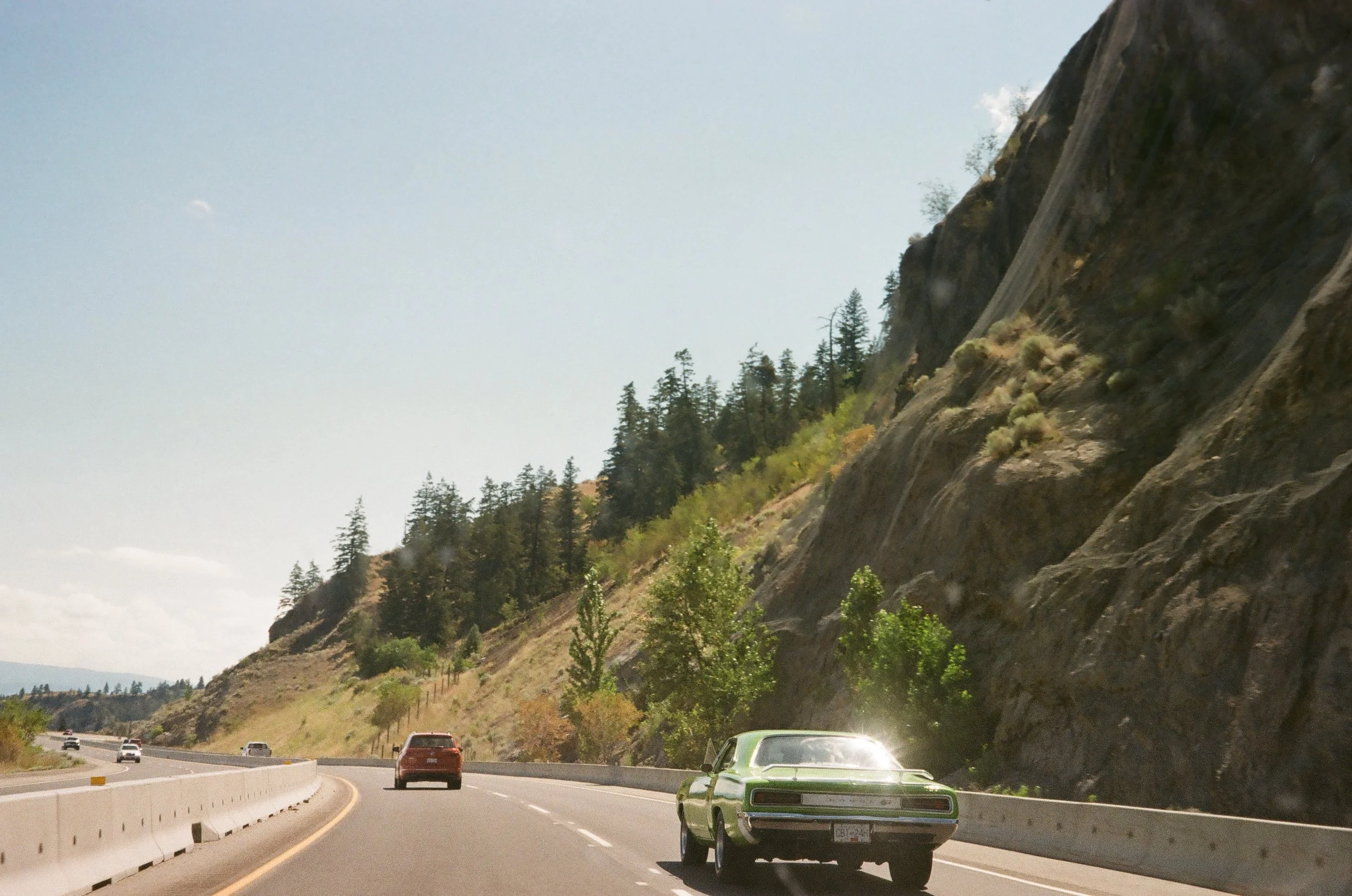 A car driving on a highway next to a rocky hillside with trees, under a partly cloudy sky.