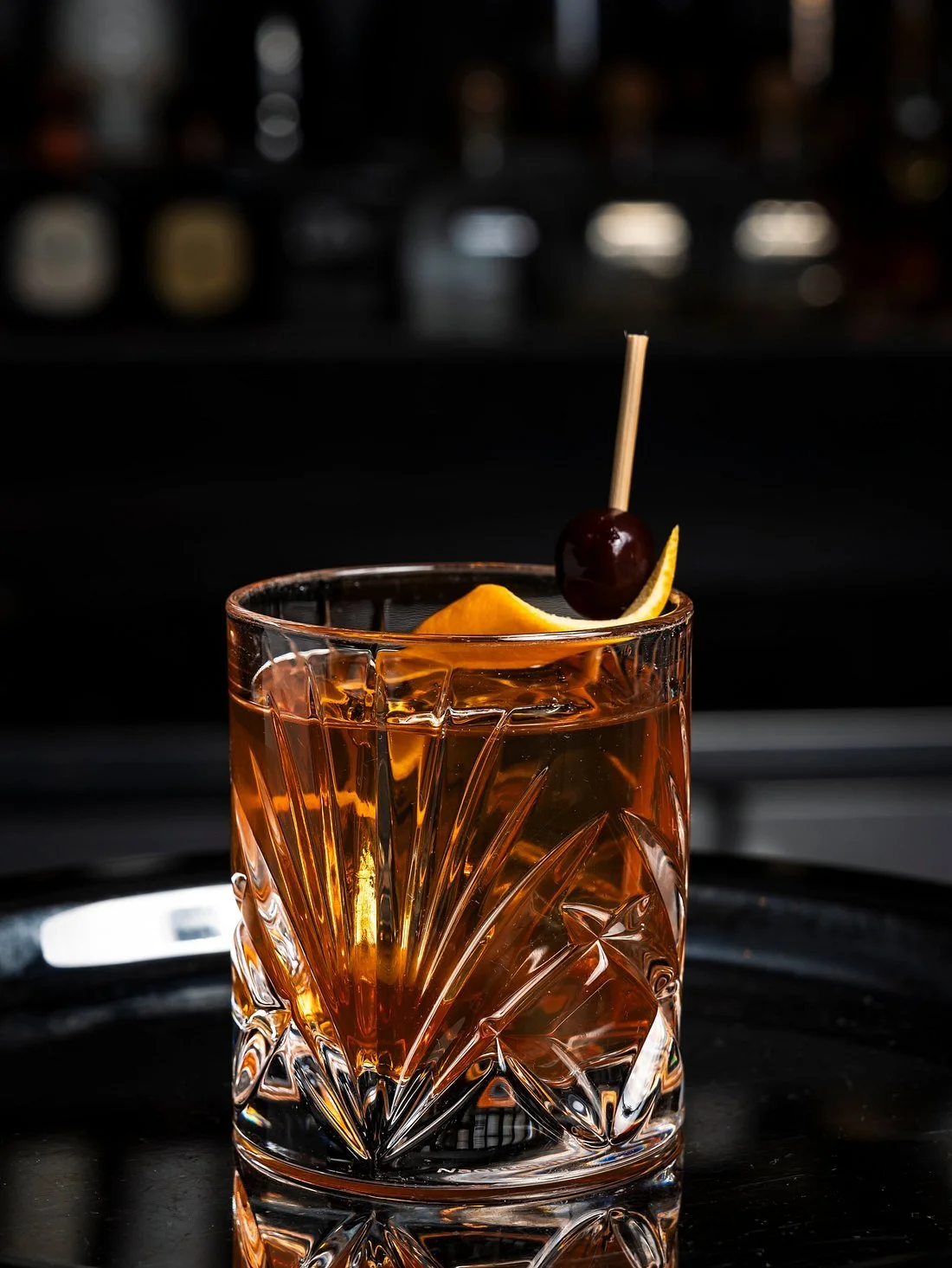 A glass of whiskey with ice, garnished with a lemon twist and a cherry on a skewer, on a dark reflective surface.