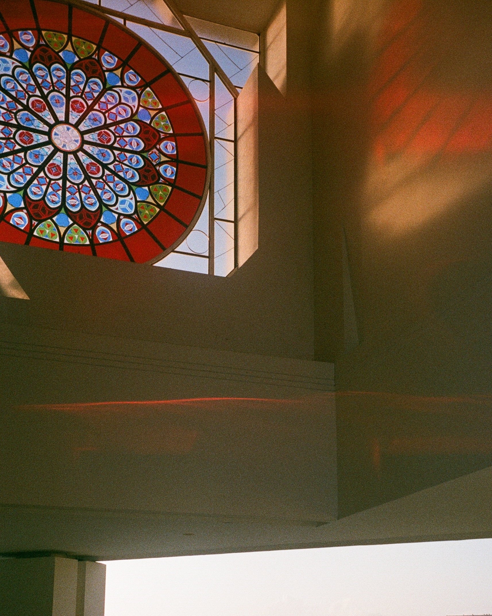 Interior view of a building with a stained glass window featuring a circular floral pattern and geometric shapes, partially visible from below.
