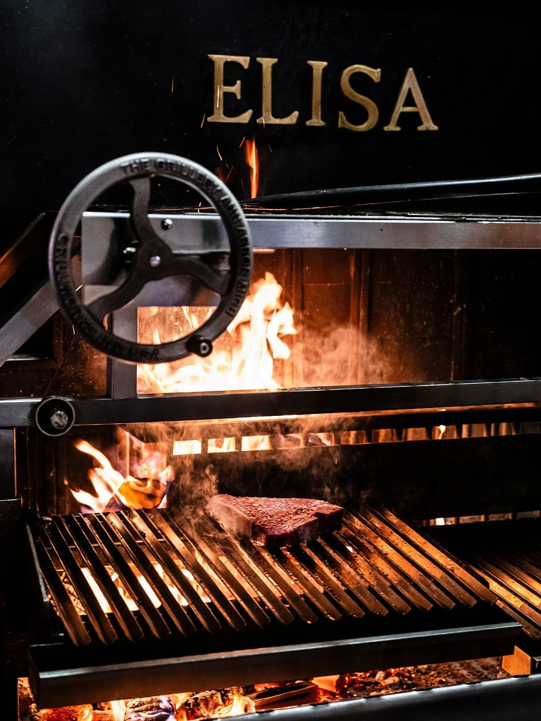 Sizzling steak on a grill with flames, under a black background with gold letters spelling 'ELISA'.