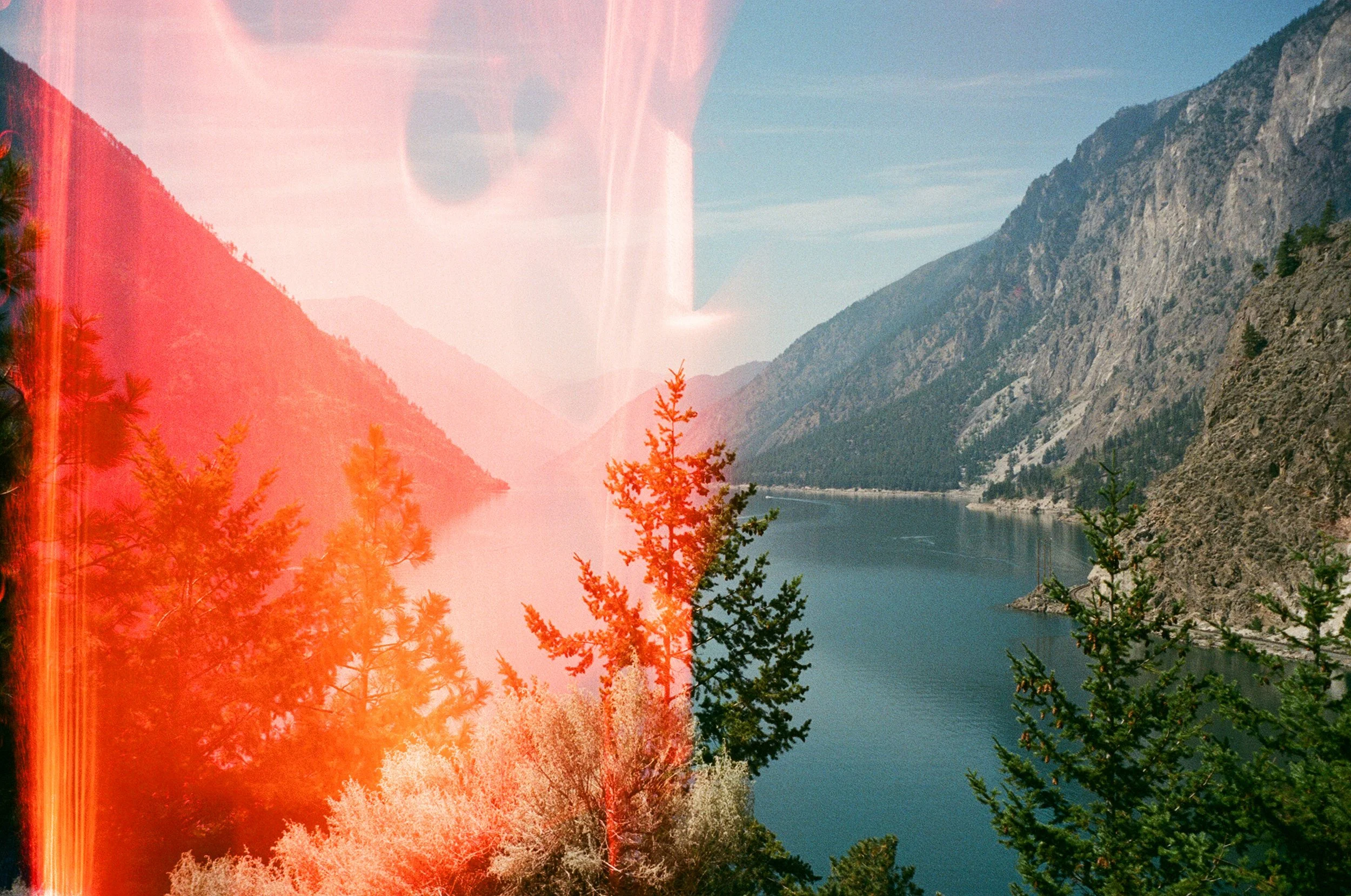 Scenic view of a river or lake surrounded by mountains with trees in the foreground. Part of the image has a red light leak or film damage effect.