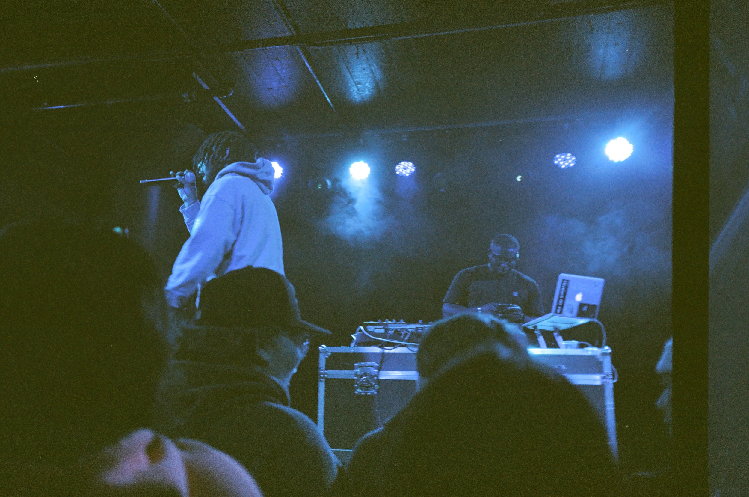 A live music performance with a vocalist singing into a microphone and a DJ in the background at a dimly lit venue with blue stage lights.