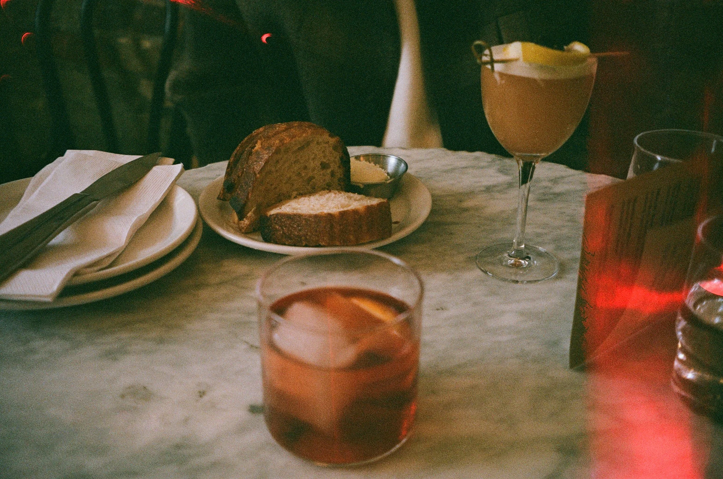 A table setting with a loaf of bread, a cold drink with lemon slices, and a cocktail glass with a lemon wedge, on a marble table in a dimly lit restaurant.