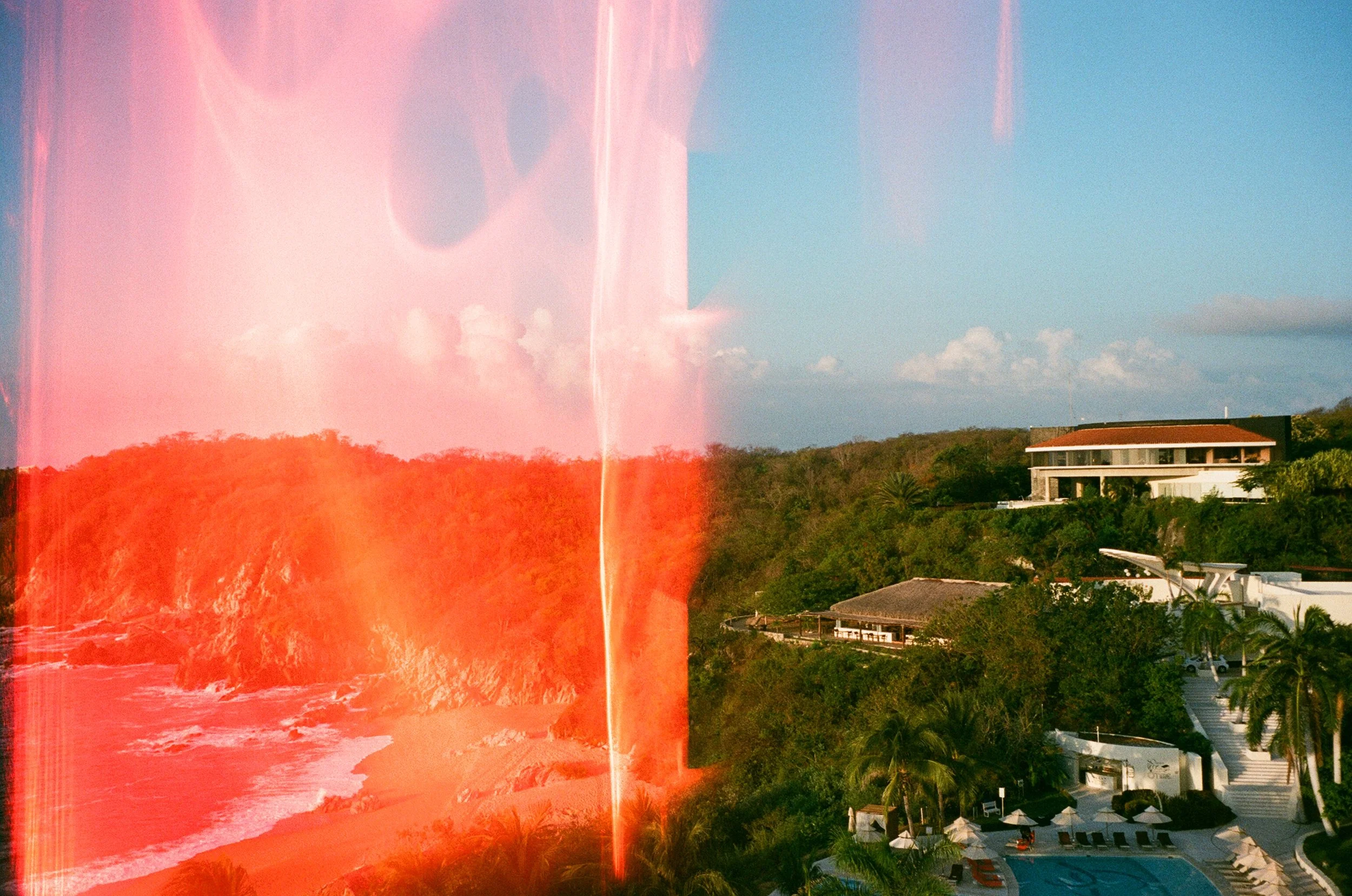 This image shows a coastal landscape with a beach, ocean waves, rocky cliffs, and lush greenery. There are buildings and a swimming pool at the bottom right, and a modern house on a hill in the background. The image has a red light leak effect on the